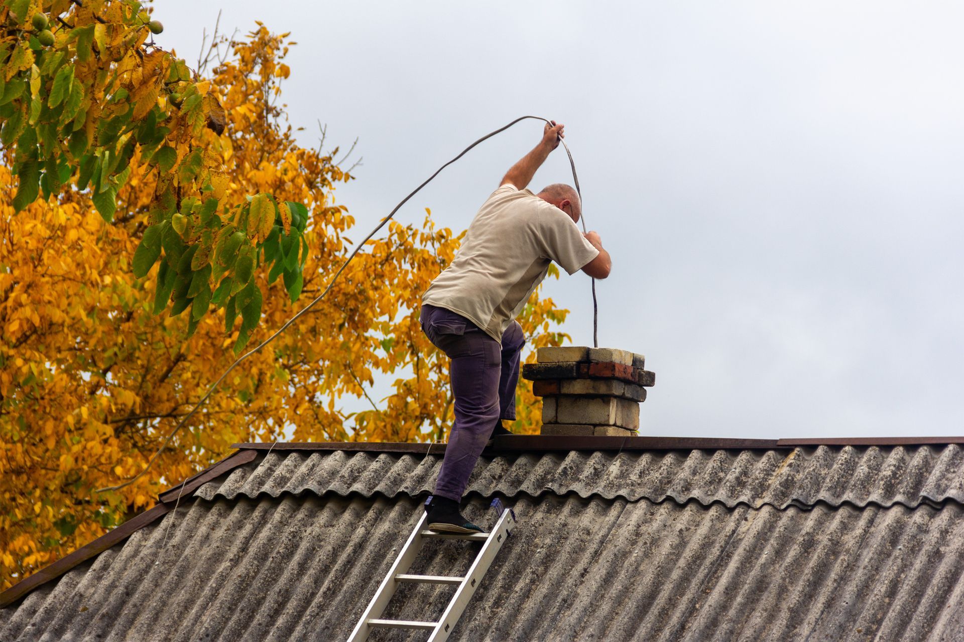 A male professional is performing chimney sweep services, from soot on the roof of a village house. 