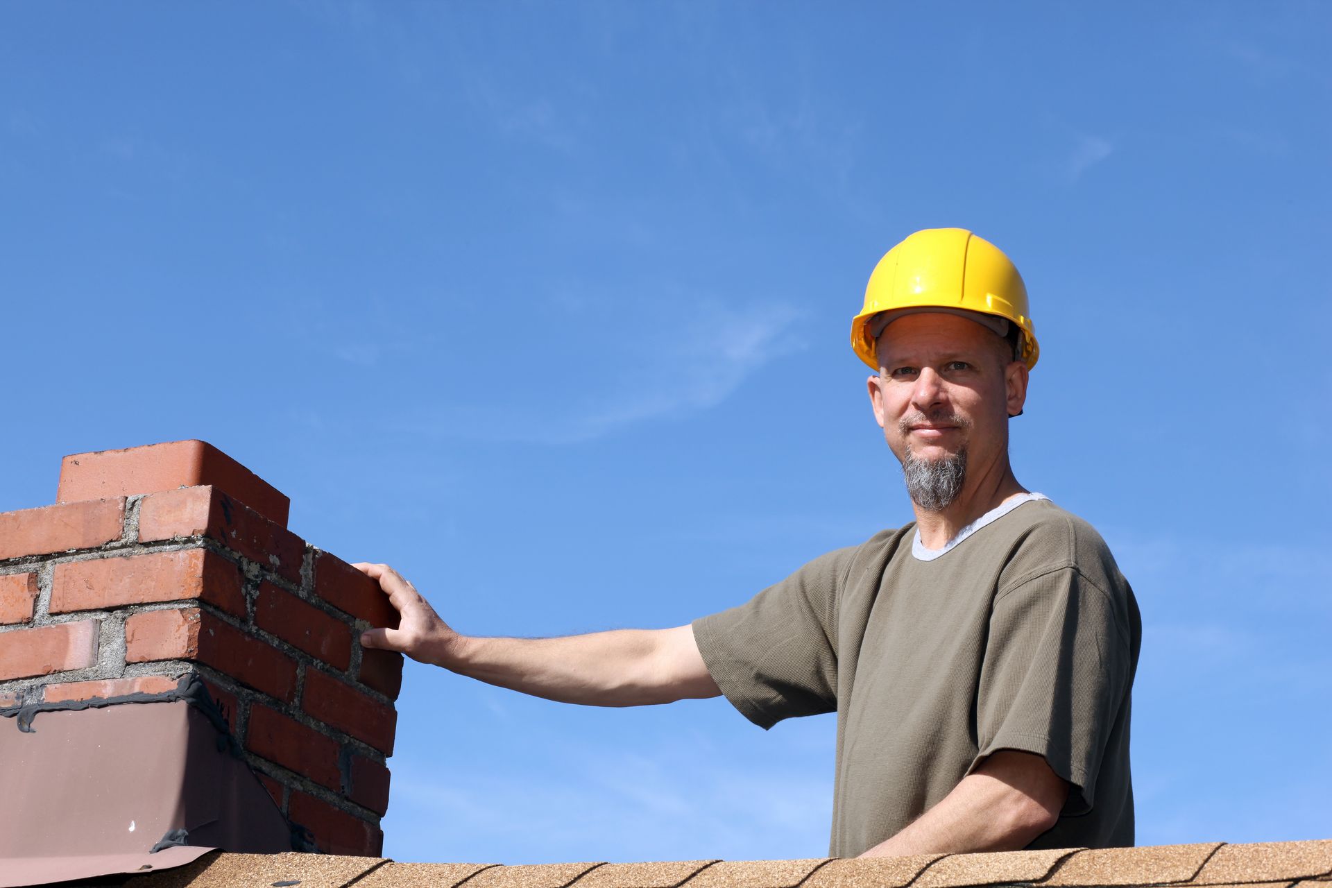 Roofing contractor in yellow hard hat inspecting brick chimney under clear blue sky.