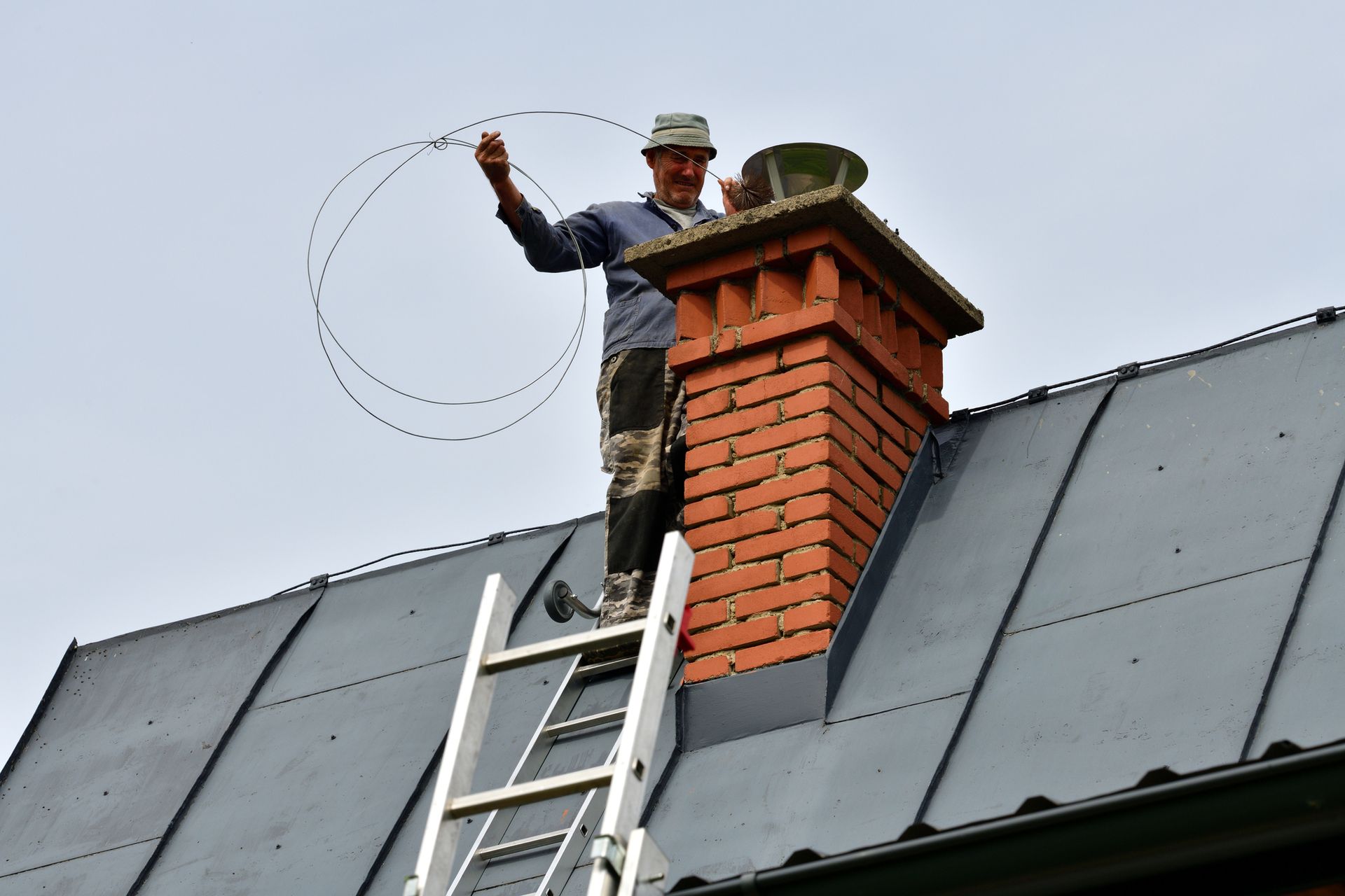 Rooftop chimney sweep serviced by technician using ladder tools.