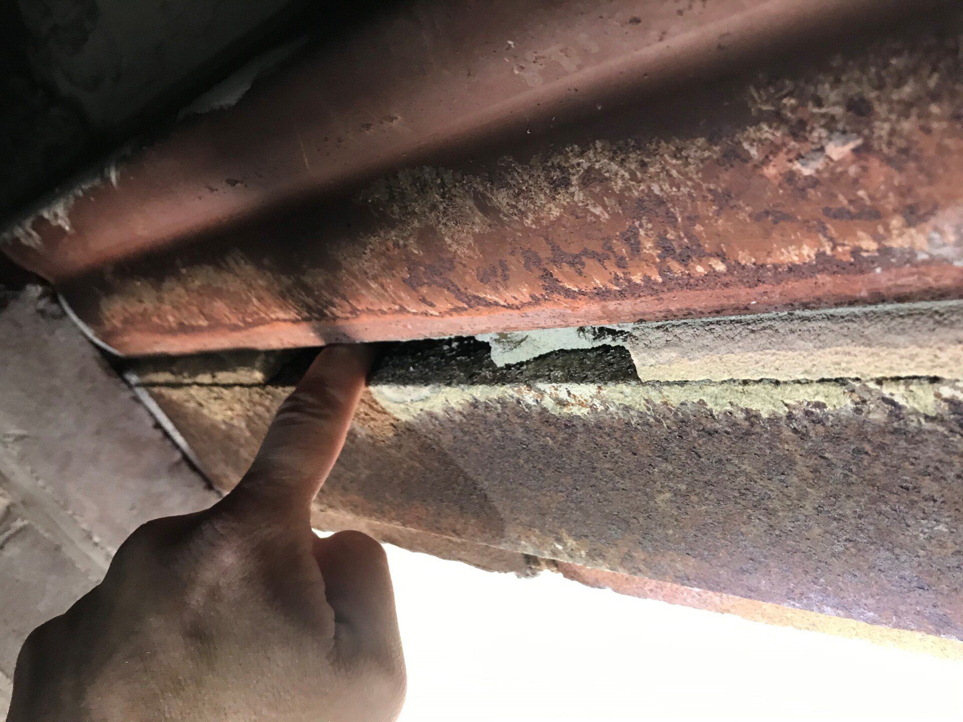 A finger points to a gap between two rusty metal panels. The bottom panel is covered in debris.