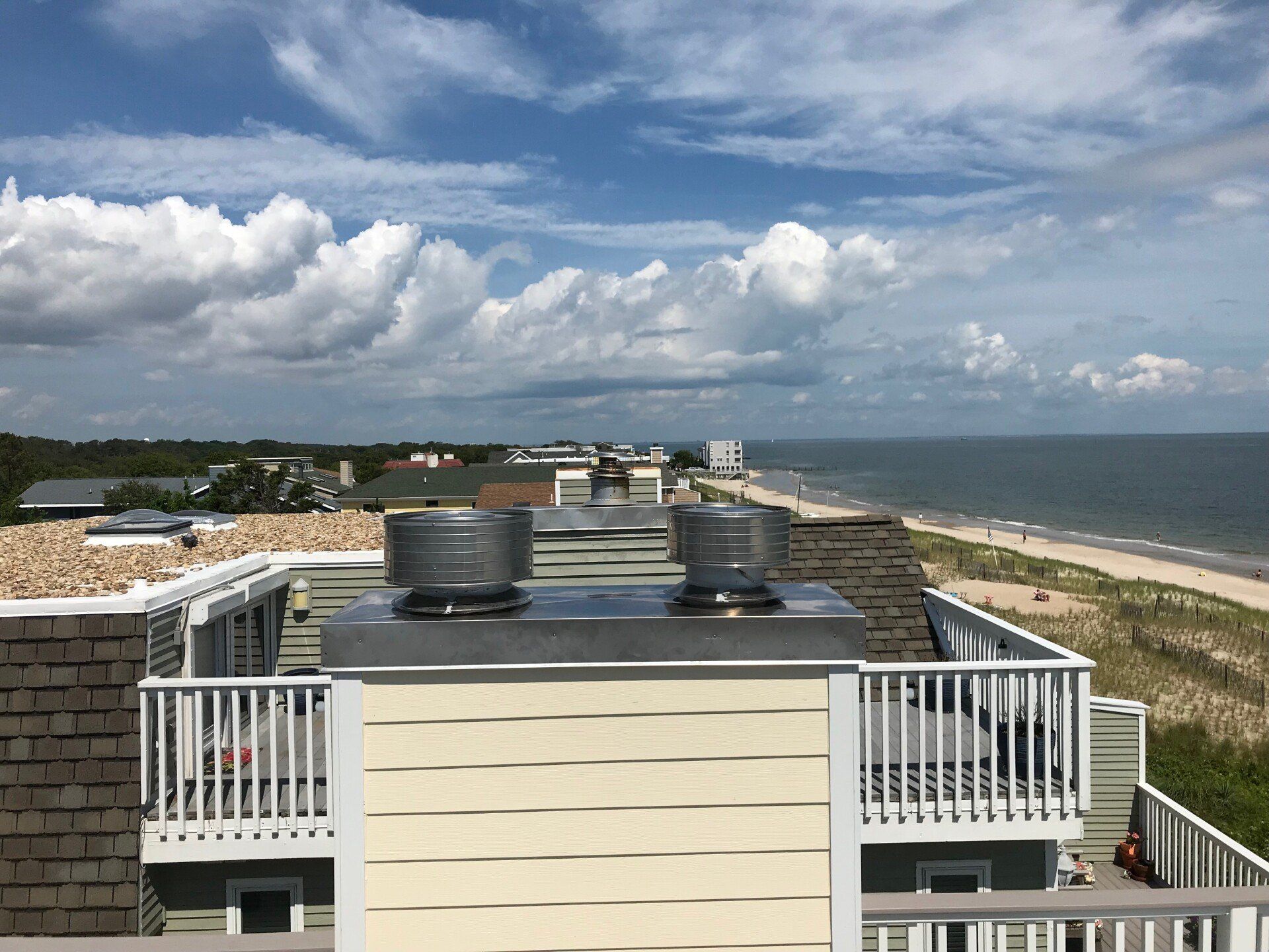 View from a rooftop overlooking the ocean. A beach and houses line the coast under a blue and cloudy sky.