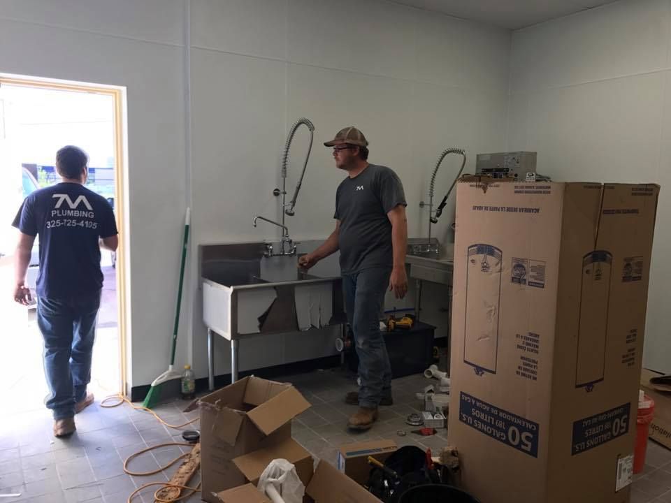Two men are standing in a room with boxes and a sink.