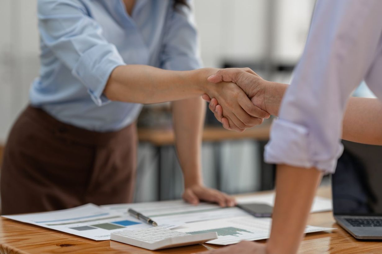 Two women shaking hands over a desk with documents, calculator, and laptop.