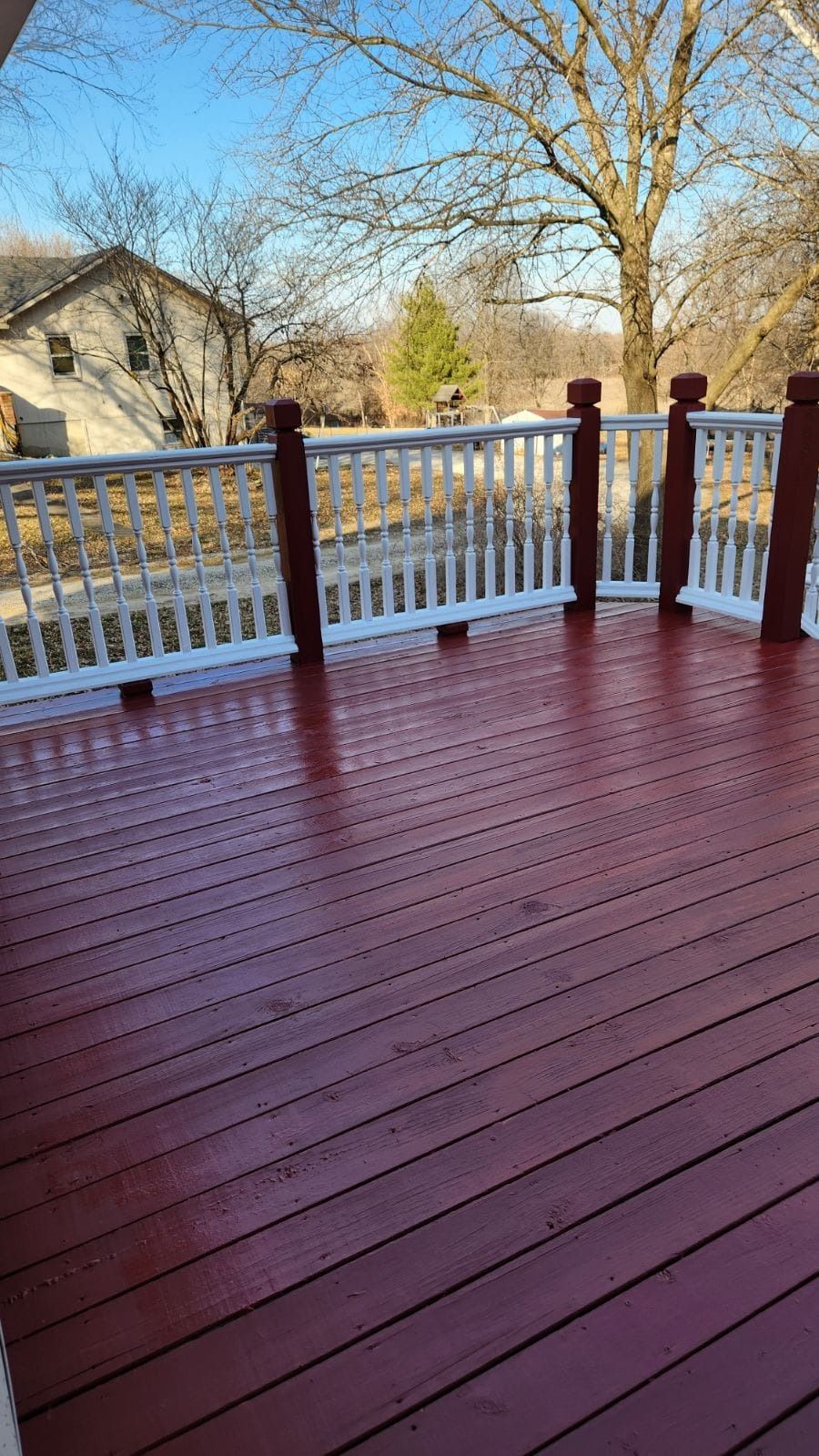 Red-stained wooden deck with white railing, and brown posts. A house and trees are in the background.