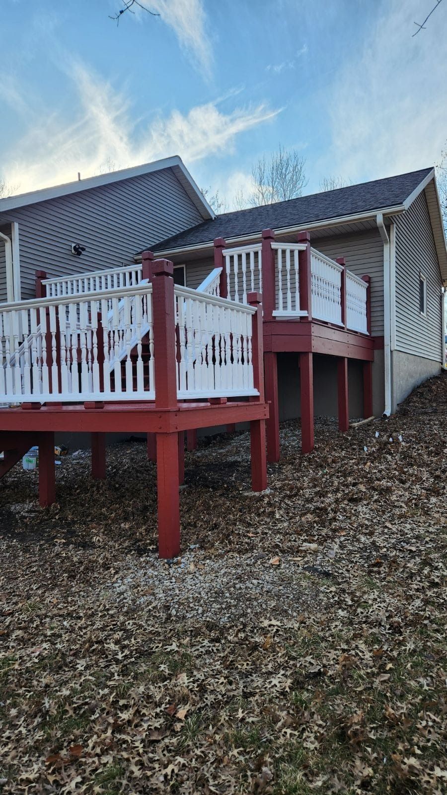 An elevated backyard deck with red support beams and white railings, attached to a house on a slope covered in dry leaves.