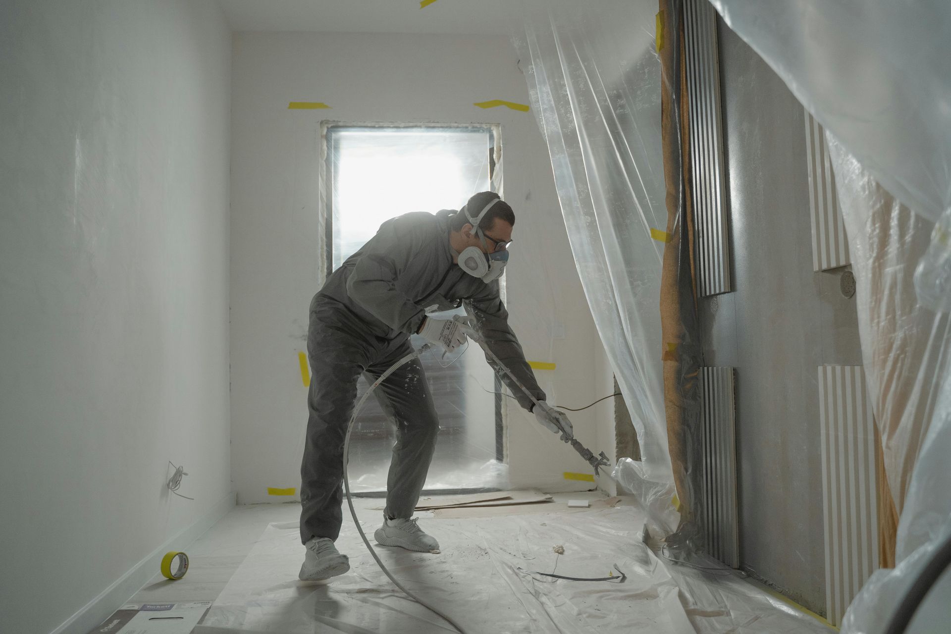 Person painting a room with a roller, wearing protective gear amid plastic-covered walls and drop cloths.