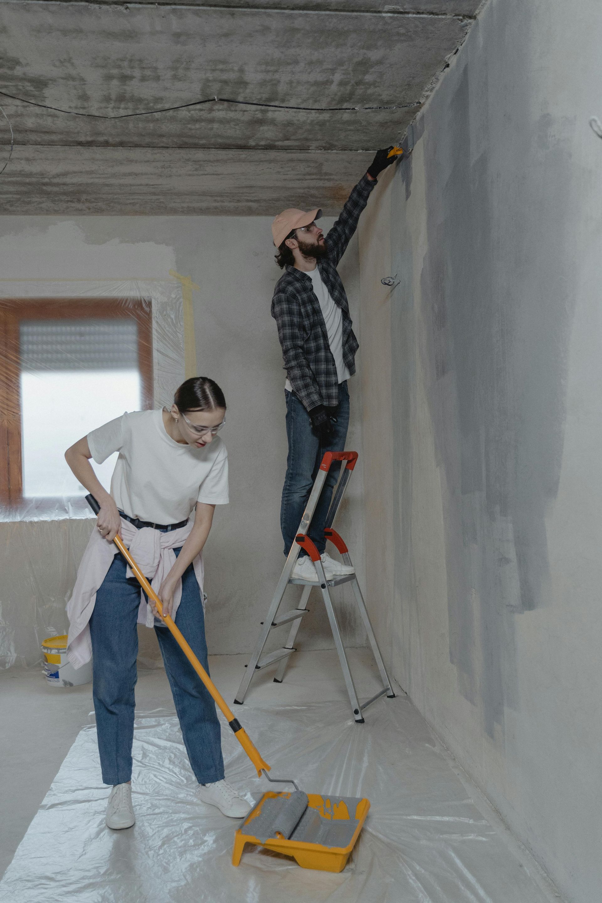 Two people painting a room: one on a ladder, one using a floor tool, with walls and floor covered for protection.