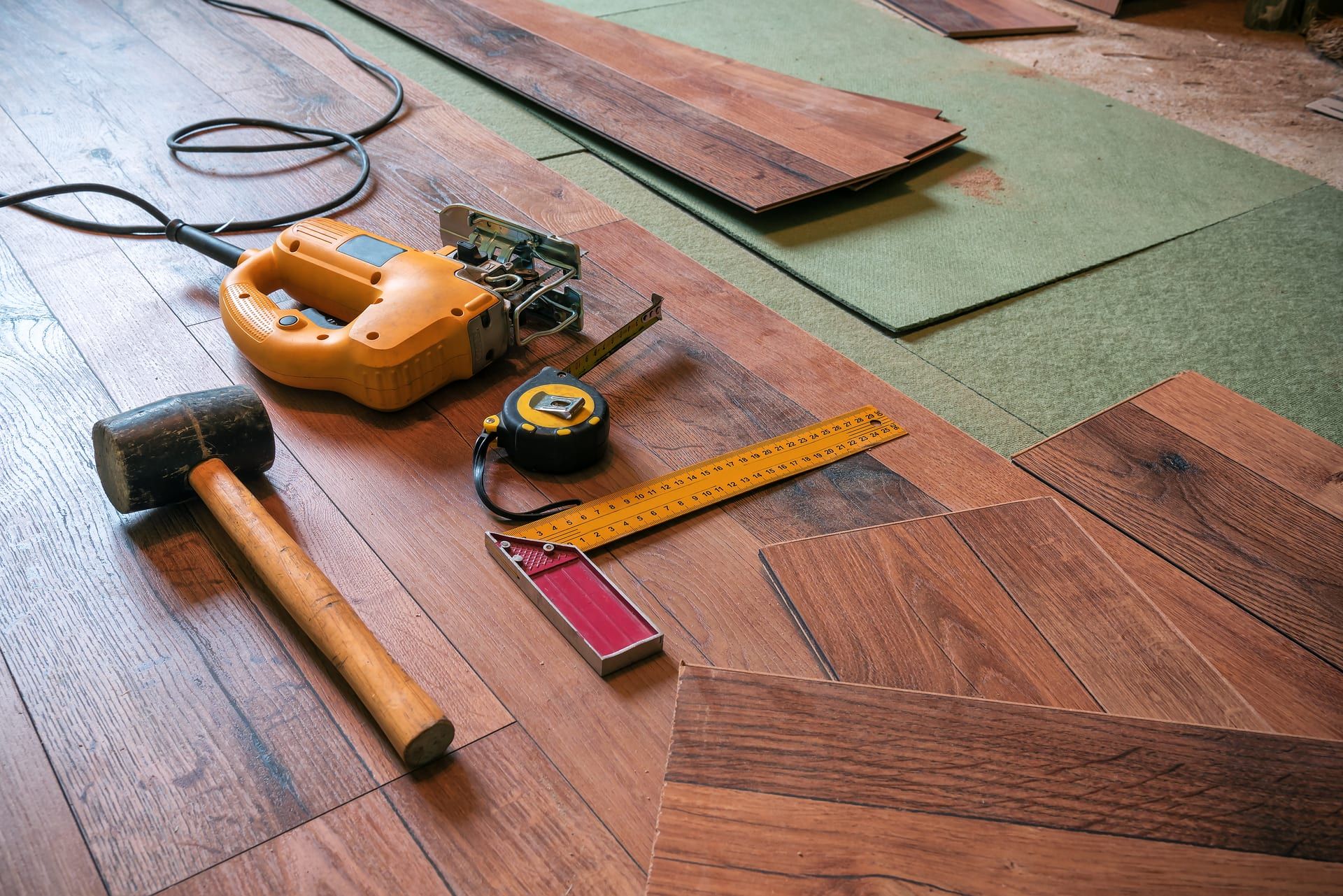 A hammer, ruler, tape measure, and saw are laying on a wooden floor.