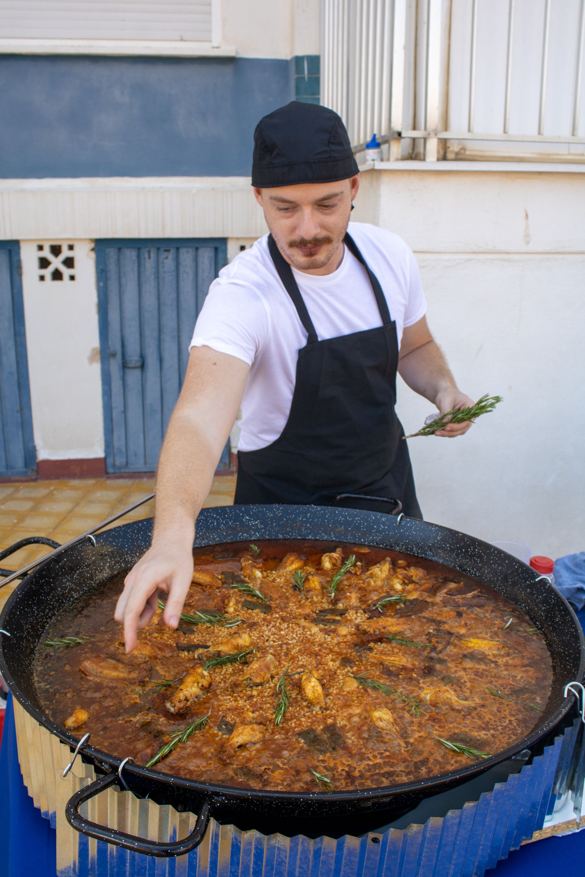 Chef adding herbs to a large paella pan, outdoors.