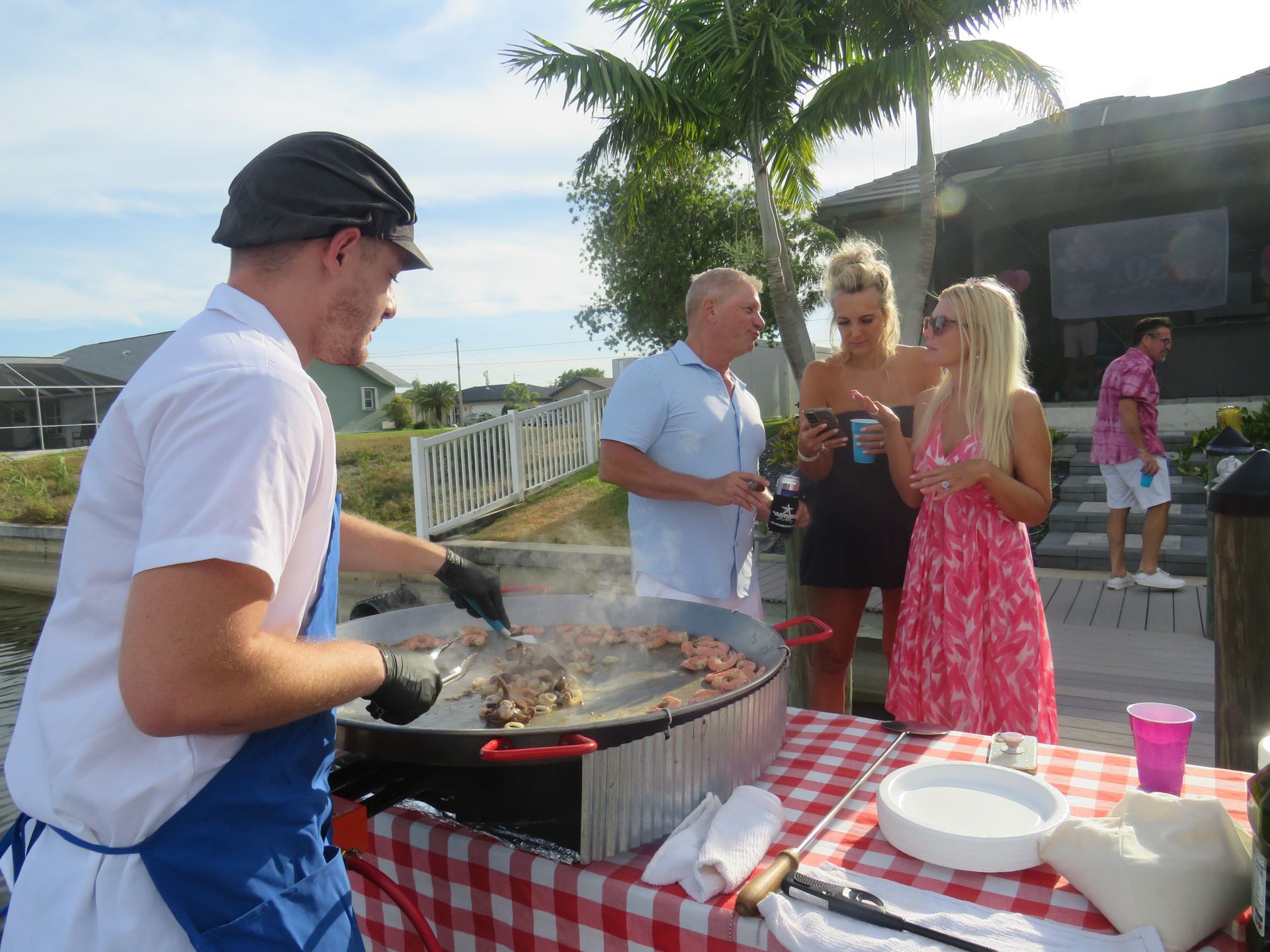Chef grilling food outdoors at a party with guests watching near a waterfront home.