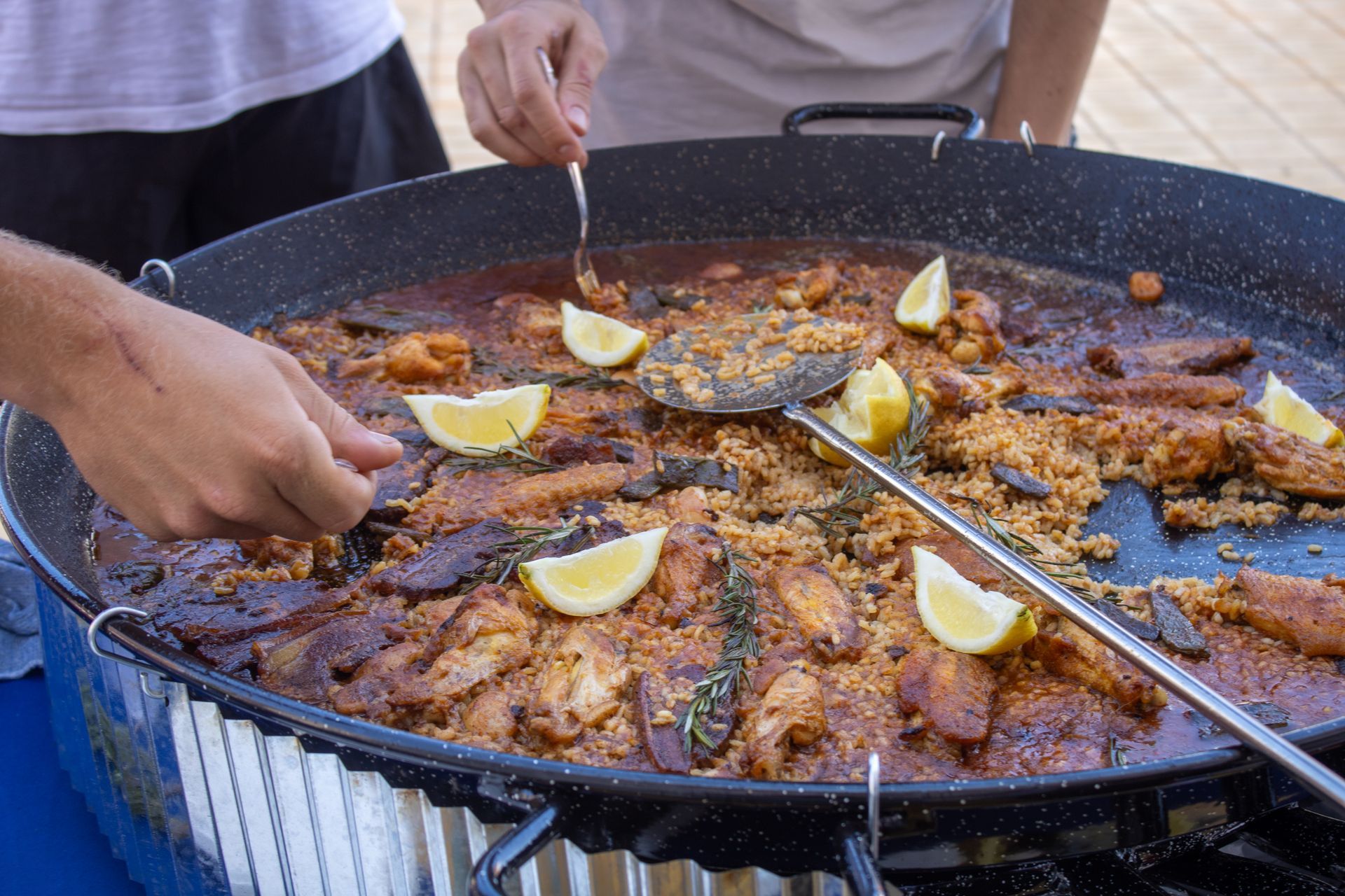 Large pan of paella, with people serving themselves.  Lemon wedges garnish the rice, seafood, and meat.