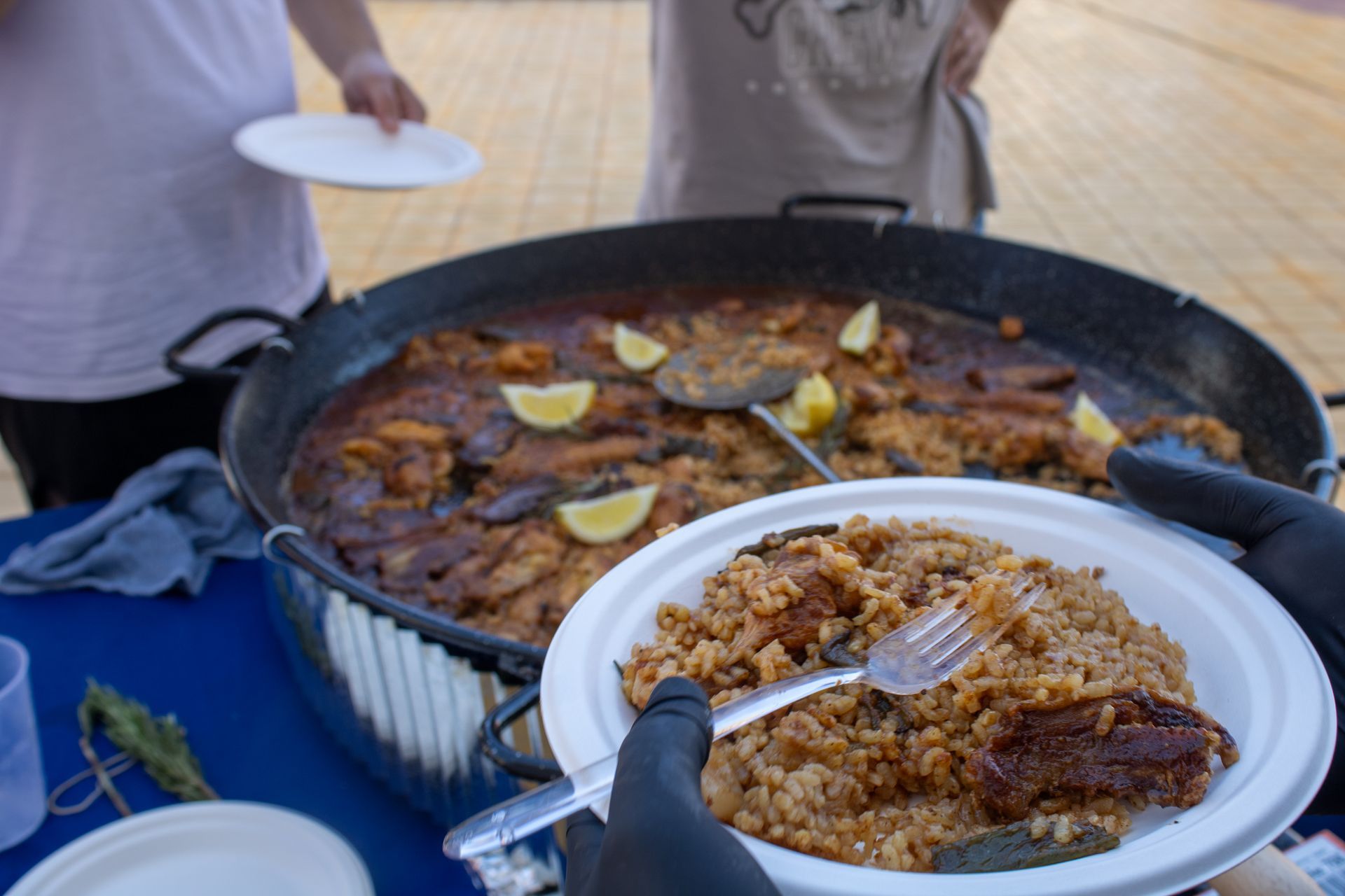 Person serving paella from a large pan onto a plate with lemon garnish, outdoors.