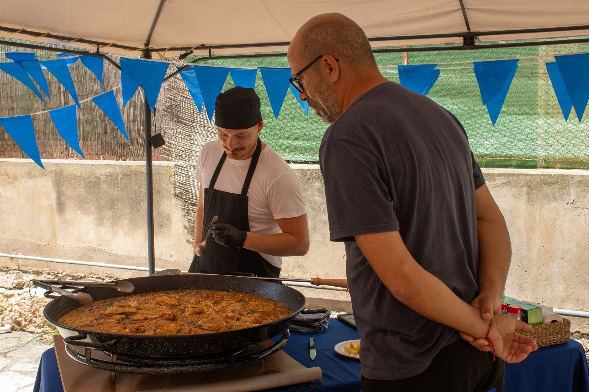 Chef in black apron serves paella to customer at a food stall, blue flags in background.
