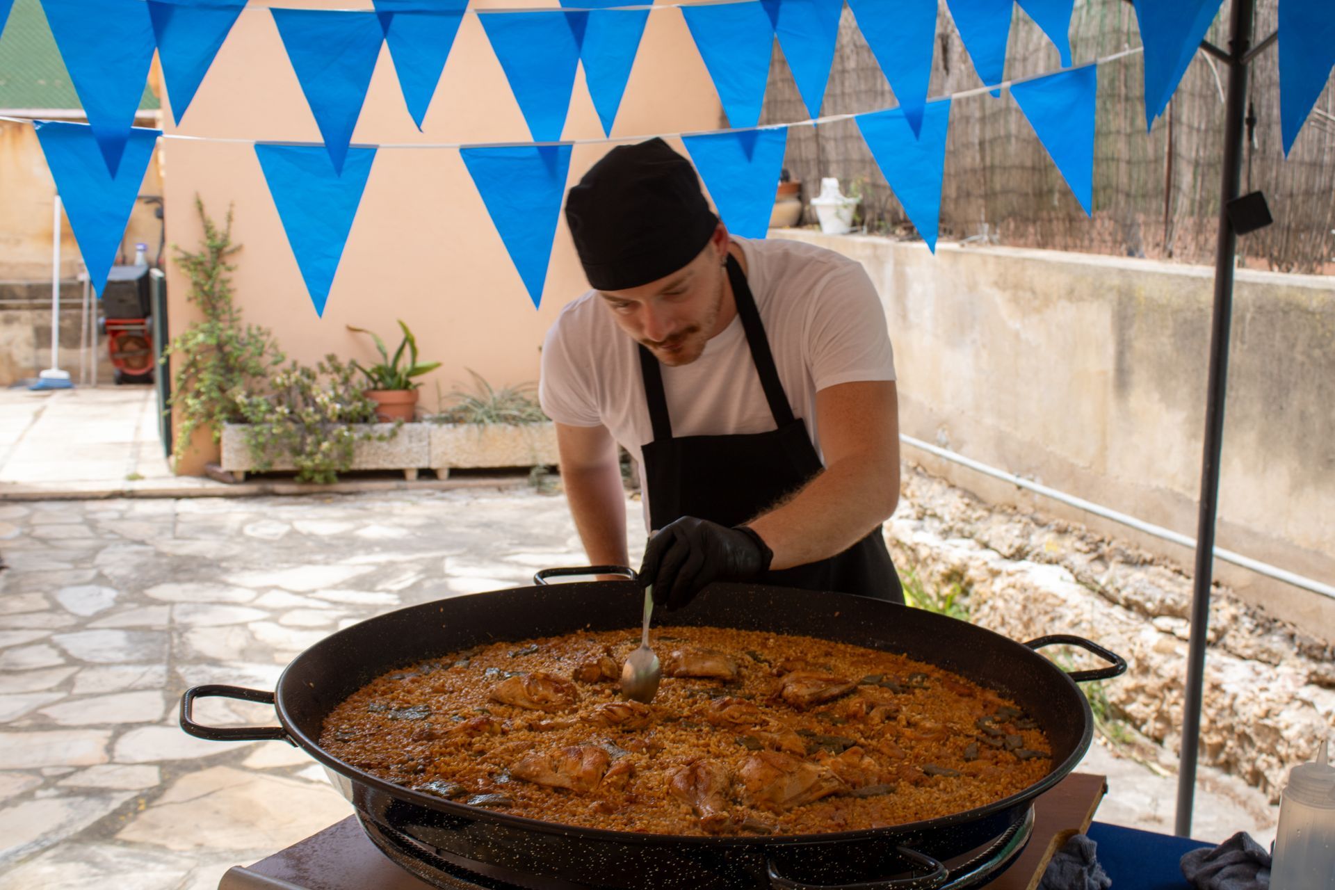 Chef in black apron and cap stirs paella in large pan, blue flags overhead.