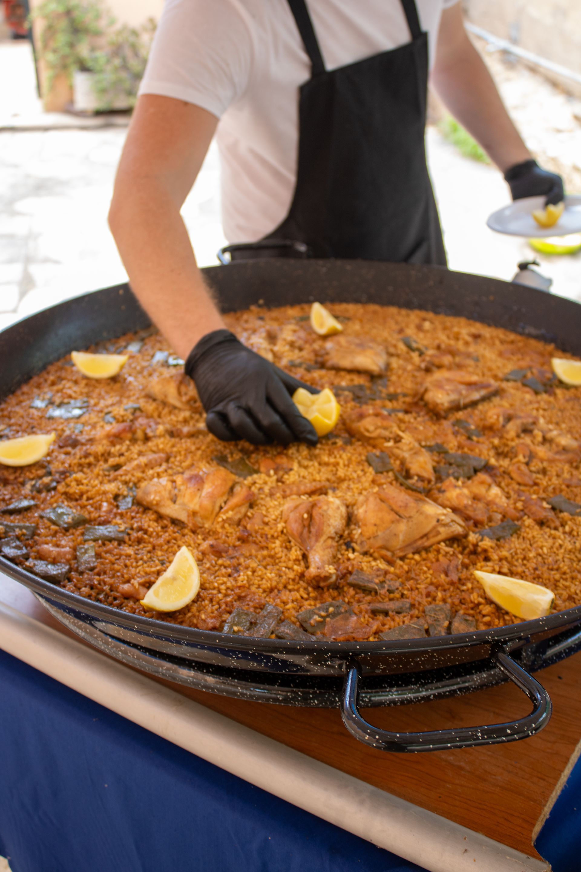 A person with black gloves garnishes a large paella with lemon wedges.