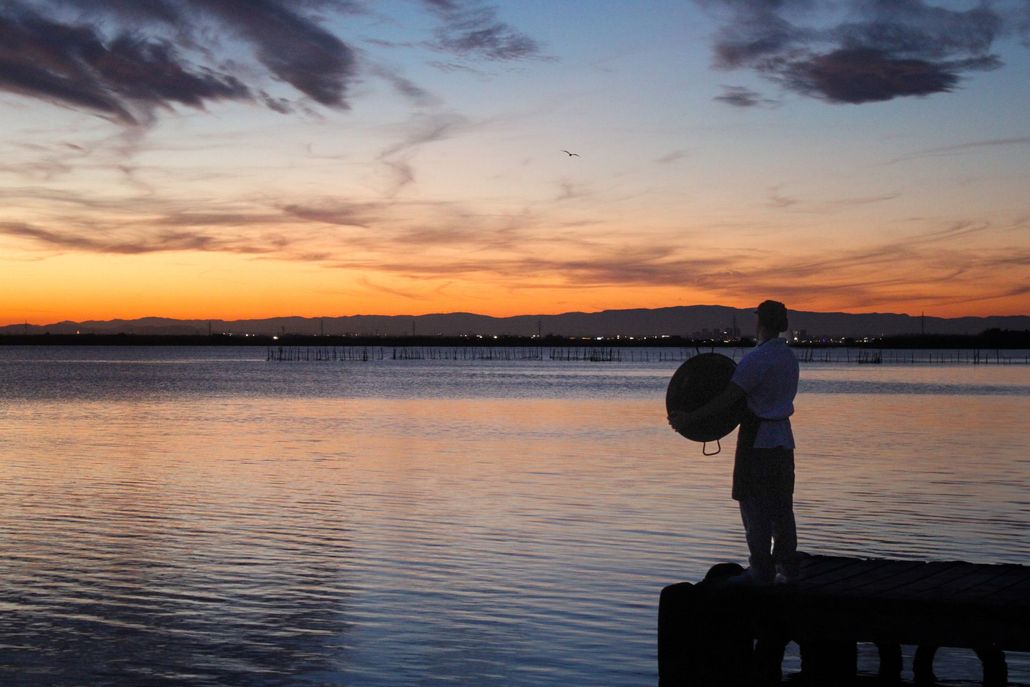 Silhouette of person holding object over water at sunset. Orange and purple sky; calm water.