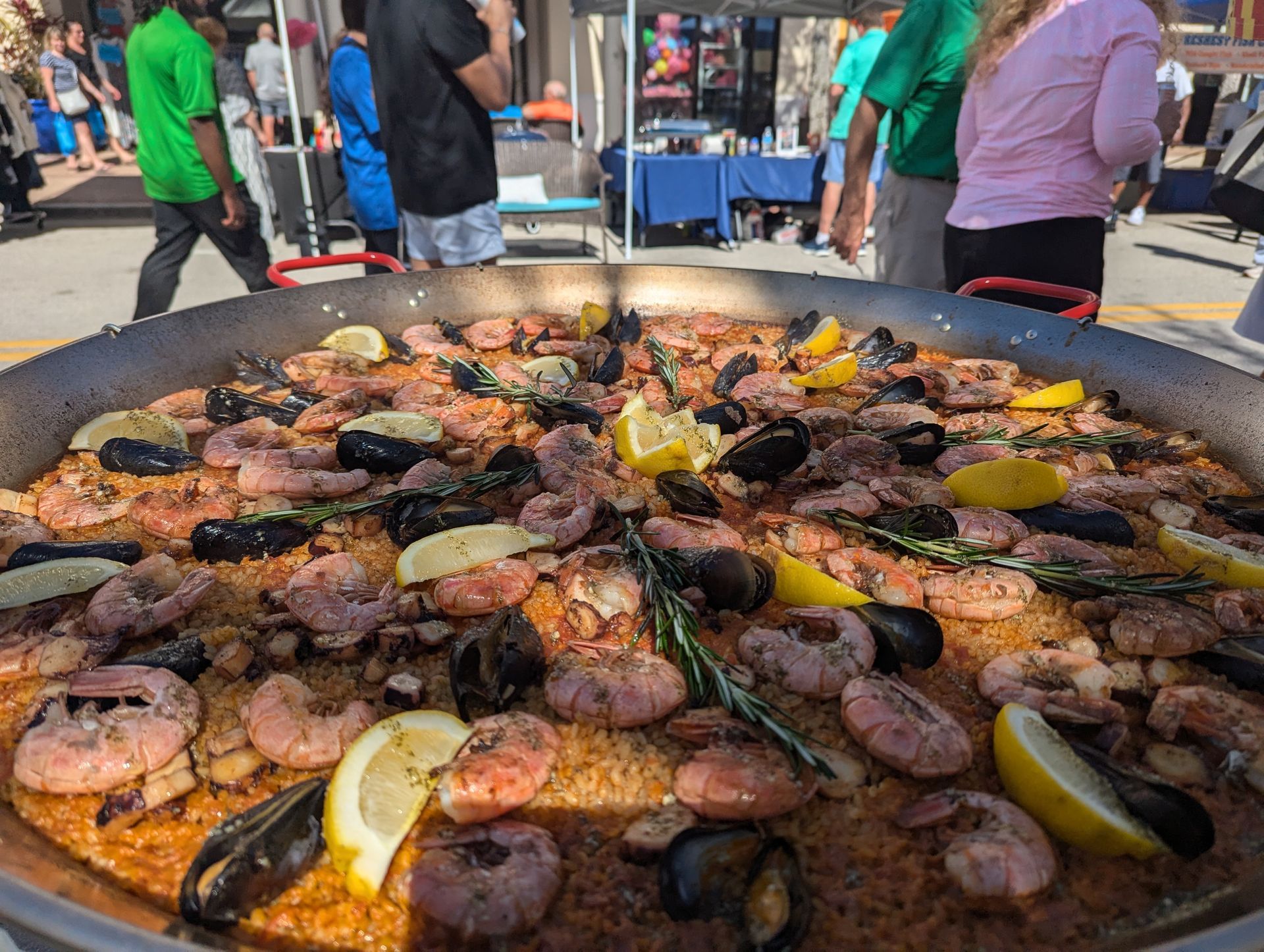 Large paella pan with seafood, lemon slices, rosemary, outdoors with people.