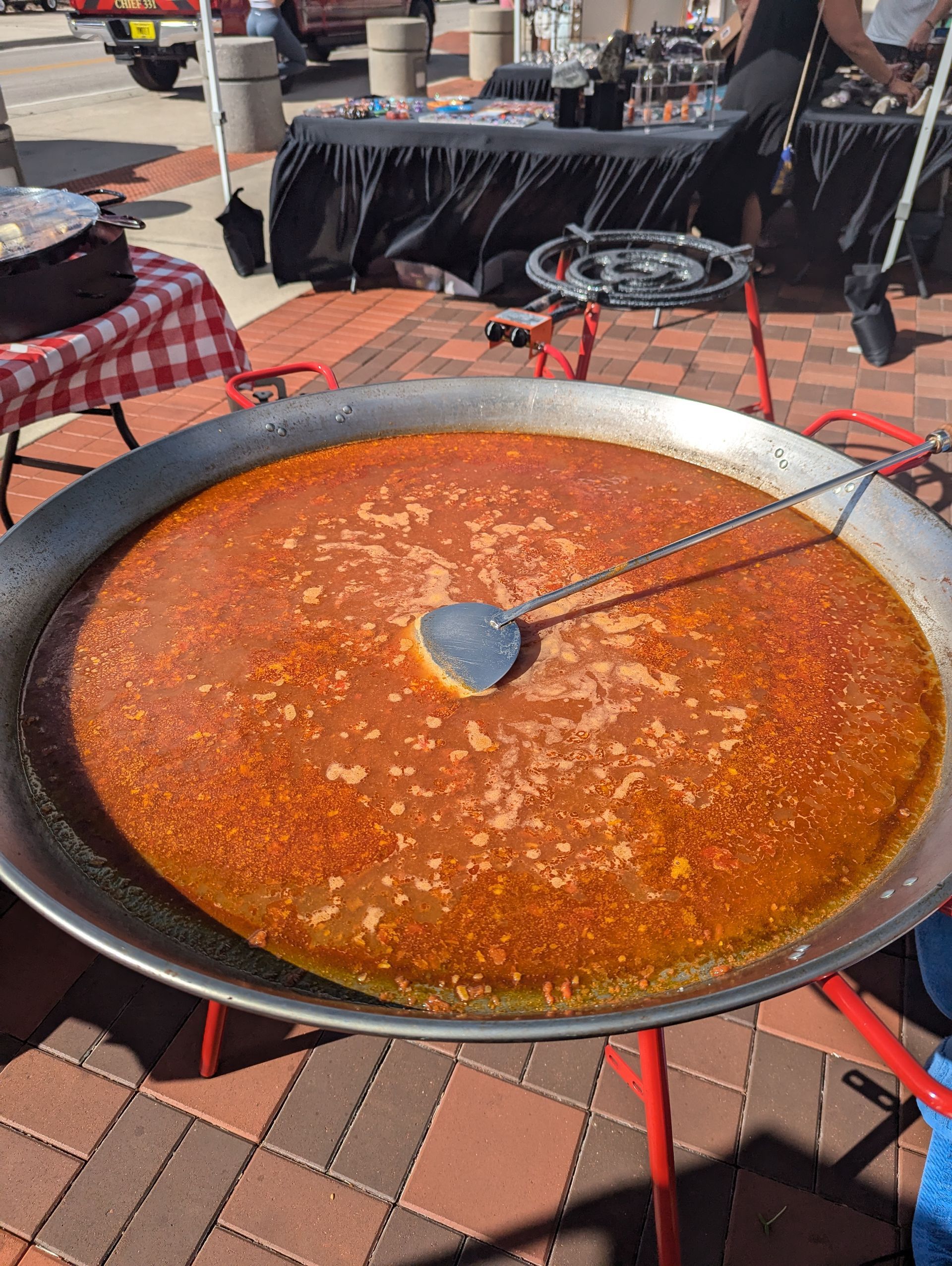 Large paella pan filled with orange rice, held by a red stand, outdoors on a brick surface.