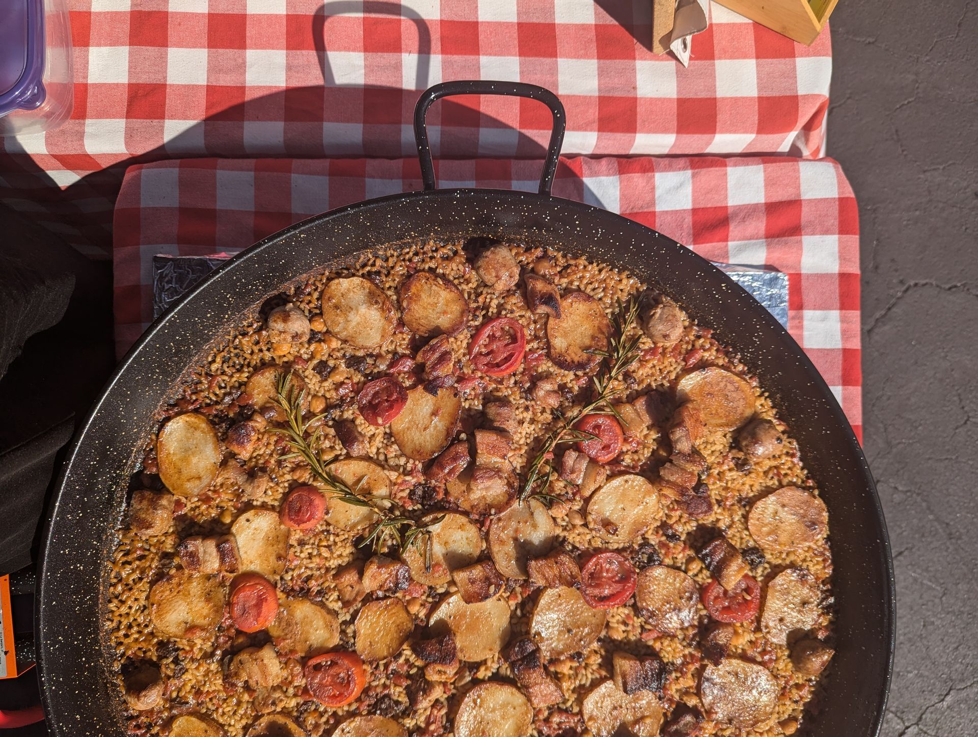 Paella in a large pan on a portable stove, topped with potatoes and tomatoes, on a red and white checkered tablecloth.