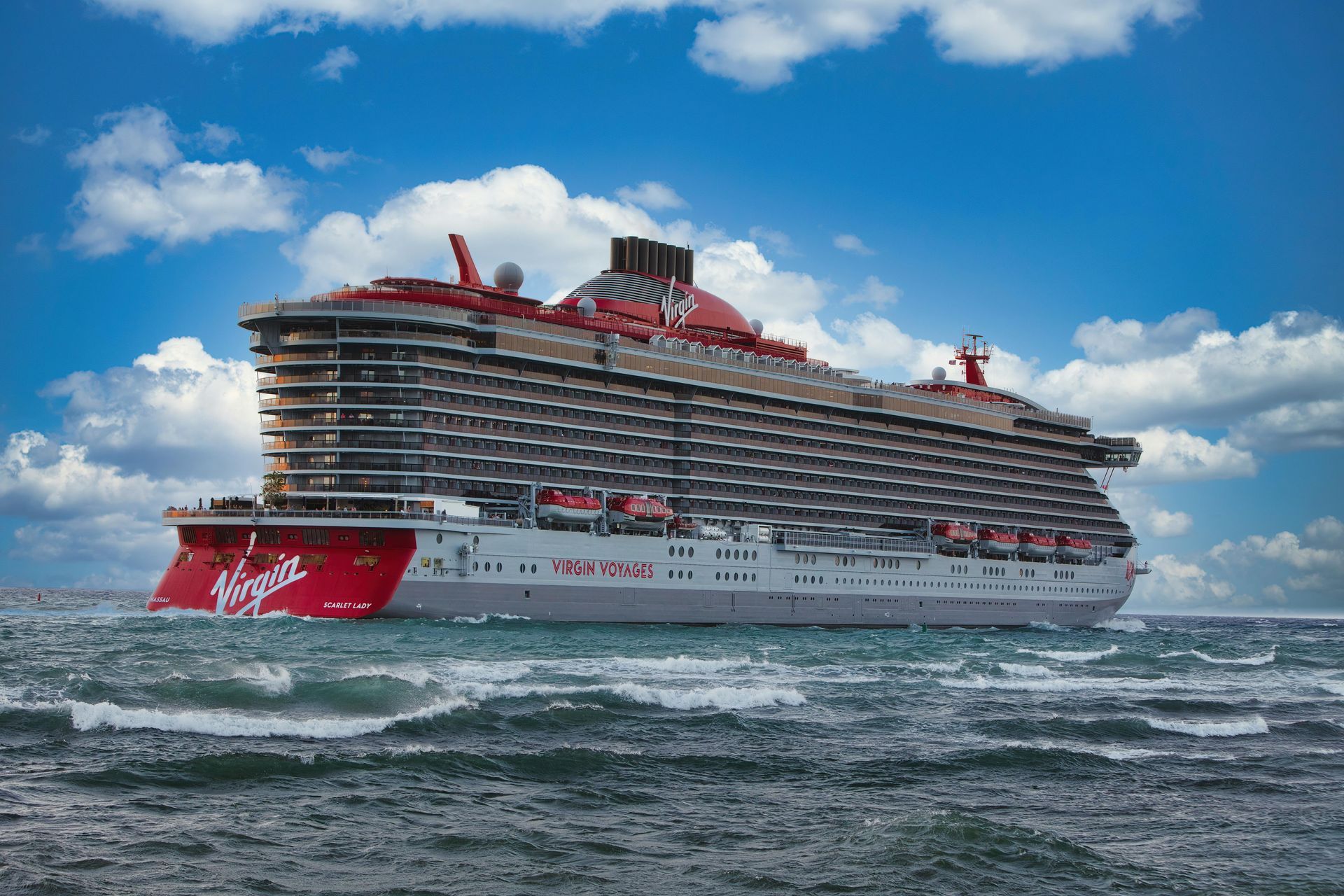 A large Virgin Voyages cruise ship on the ocean, red and gray, with a blue sky background.