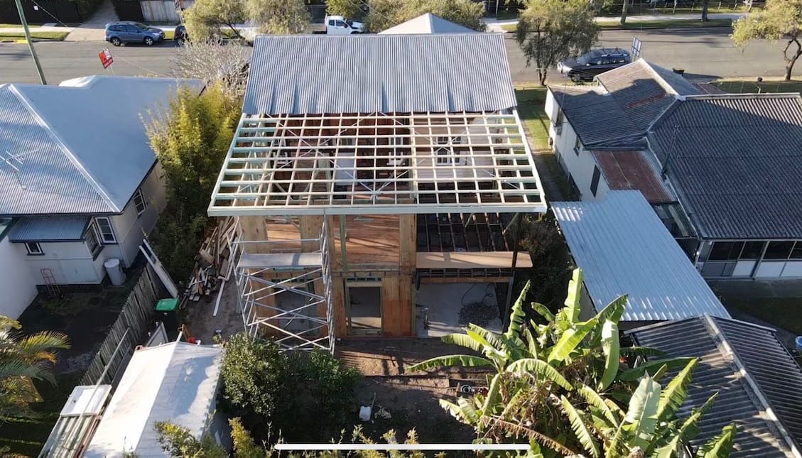 Aerial view of a two-story house under construction. Unfinished metal roof, wood framing, and surrounding houses.