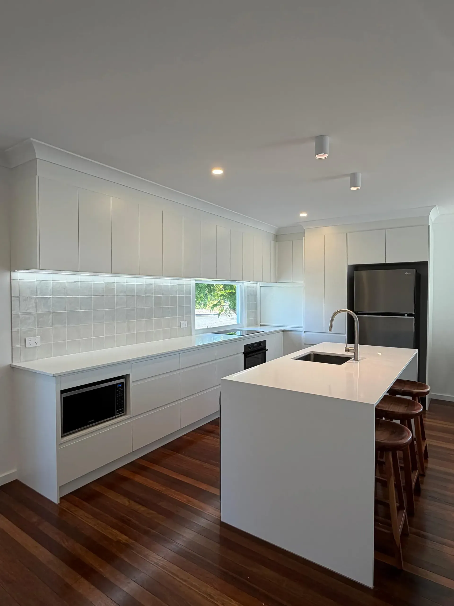 Modern white kitchen with island, wooden floor, and stainless steel refrigerator.