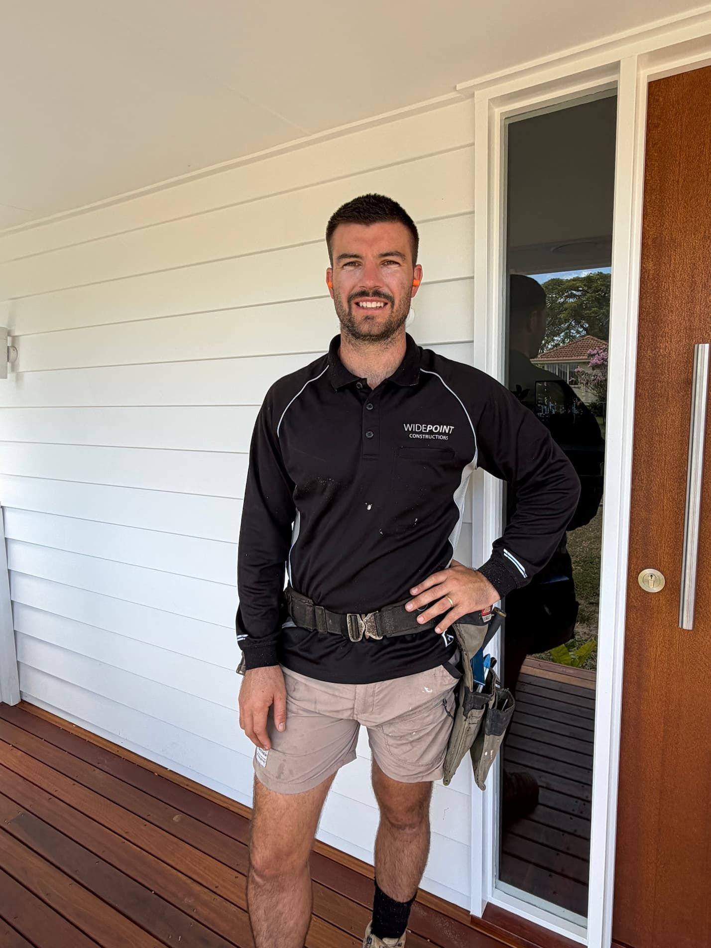 Man in work clothes poses on a porch, smiling. He wears a black shirt, tan shorts, and a tool belt.
