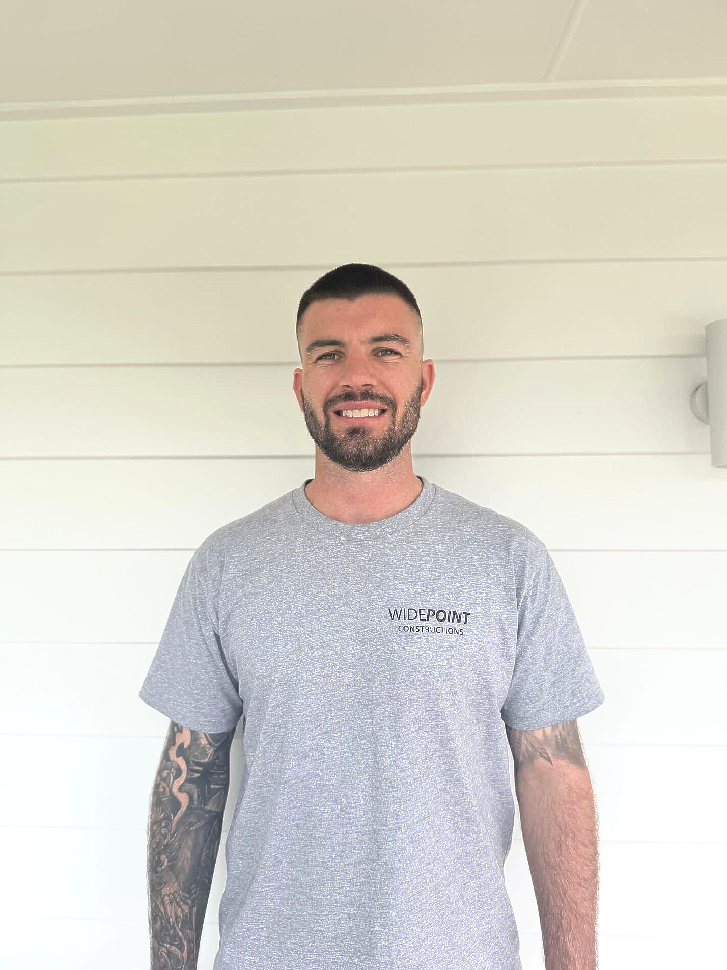 Man in work clothes standing on a porch, smiling. He's wearing shorts and a tool belt.