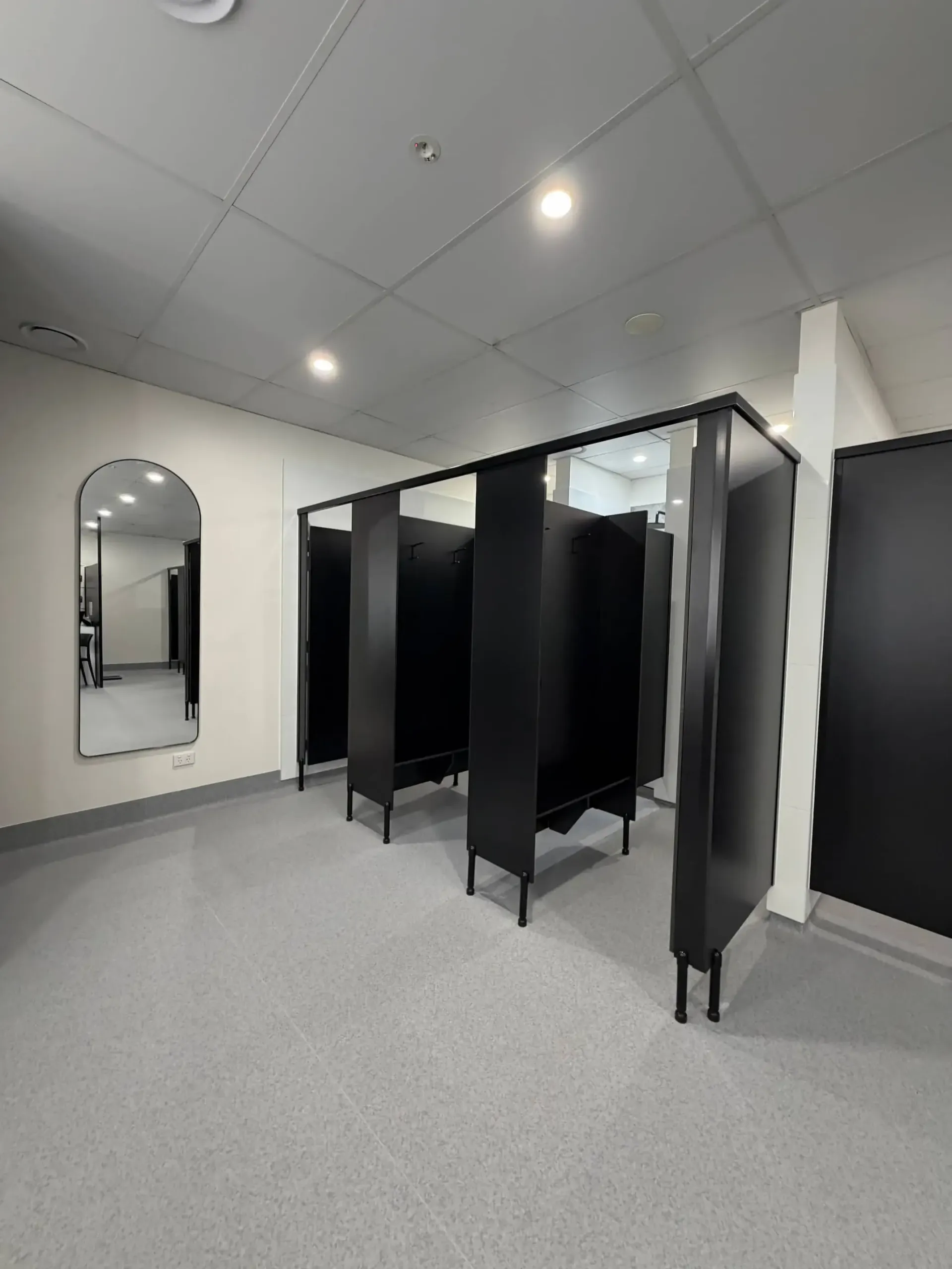 Black restroom stalls with gray flooring and a large arched mirror.