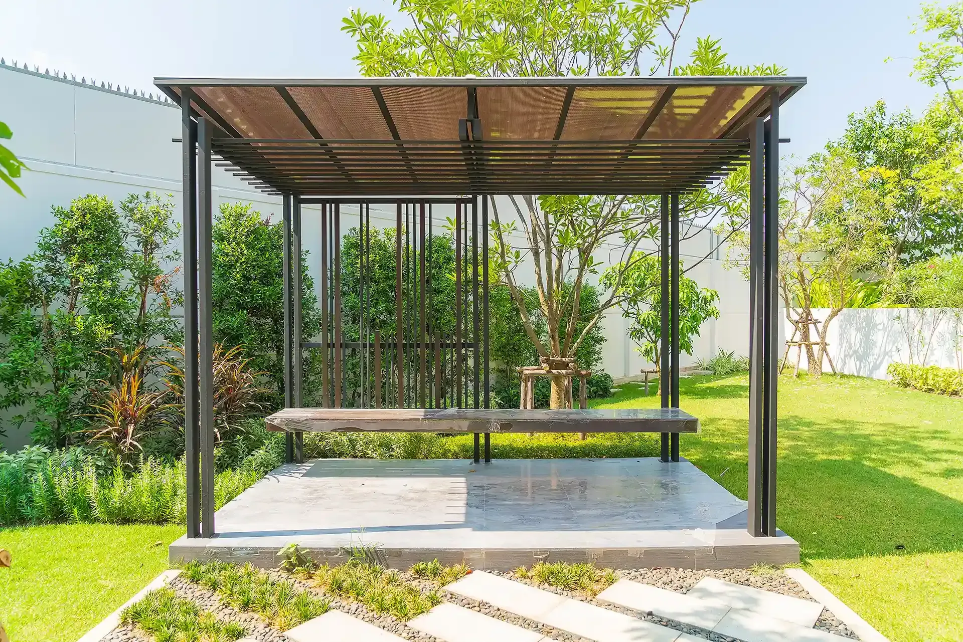Gazebo in a sunny garden with a bench, metal frame, and shaded roof.