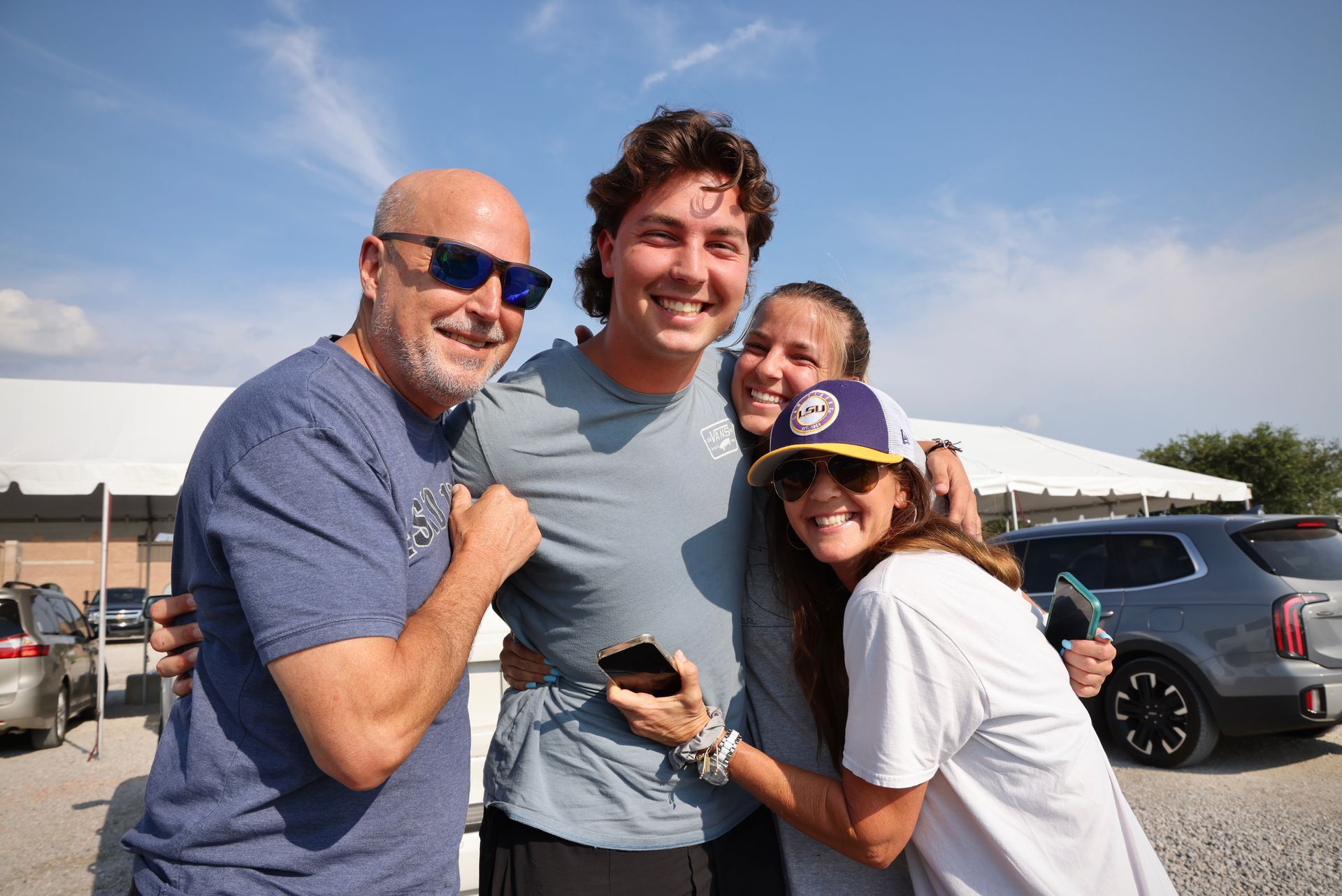 Three smiling people pose outdoors in front of parked cars on a sunny day.