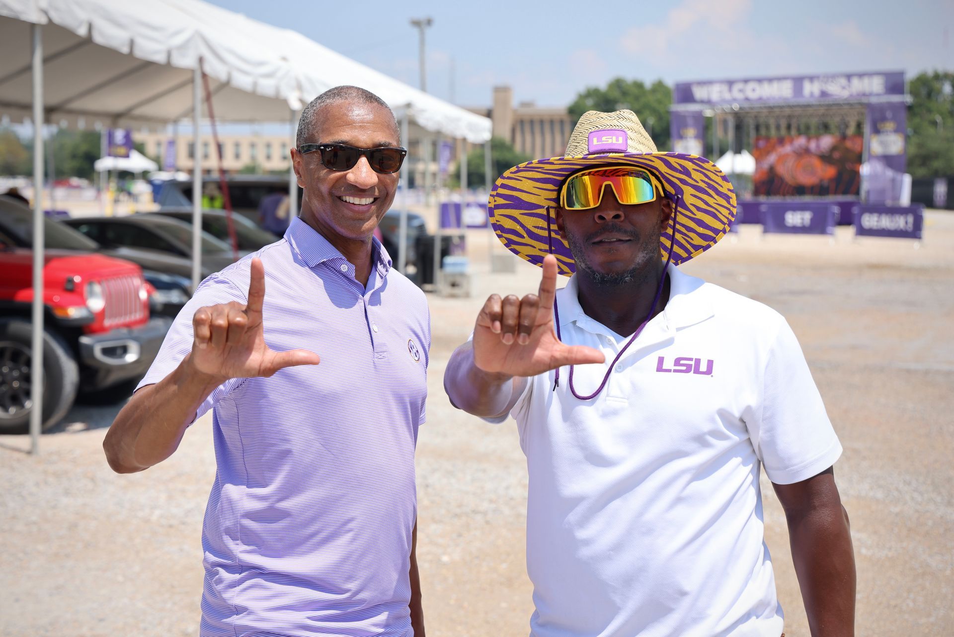 Two men posing outdoors, smiling and making hand signs at a car show entrance