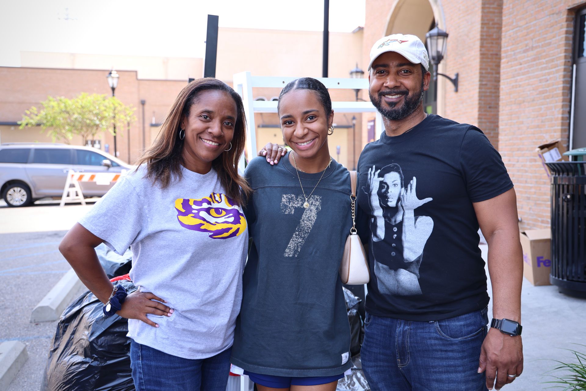 Three smiling people posing outdoors, two in T-shirts and one in a black shirt and white cap.