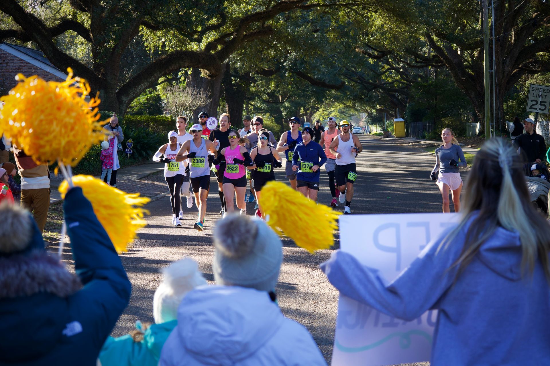 Runners in a park race past cheering spectators waving bright yellow pom-poms.