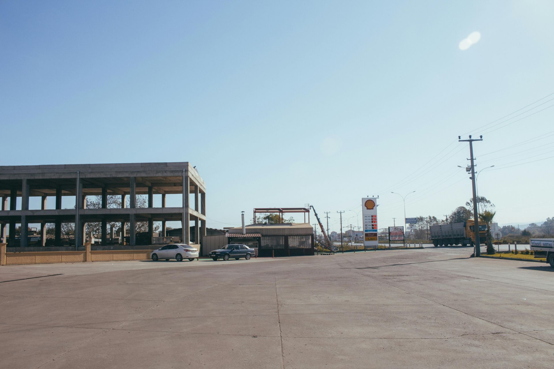Empty lot with a partially built concrete building, a small kiosk, and a gas station sign under a blue sky.