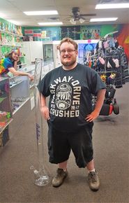 Man with a giant bong, standing in a smoke shop. Woman in tie-dye smiles in background.