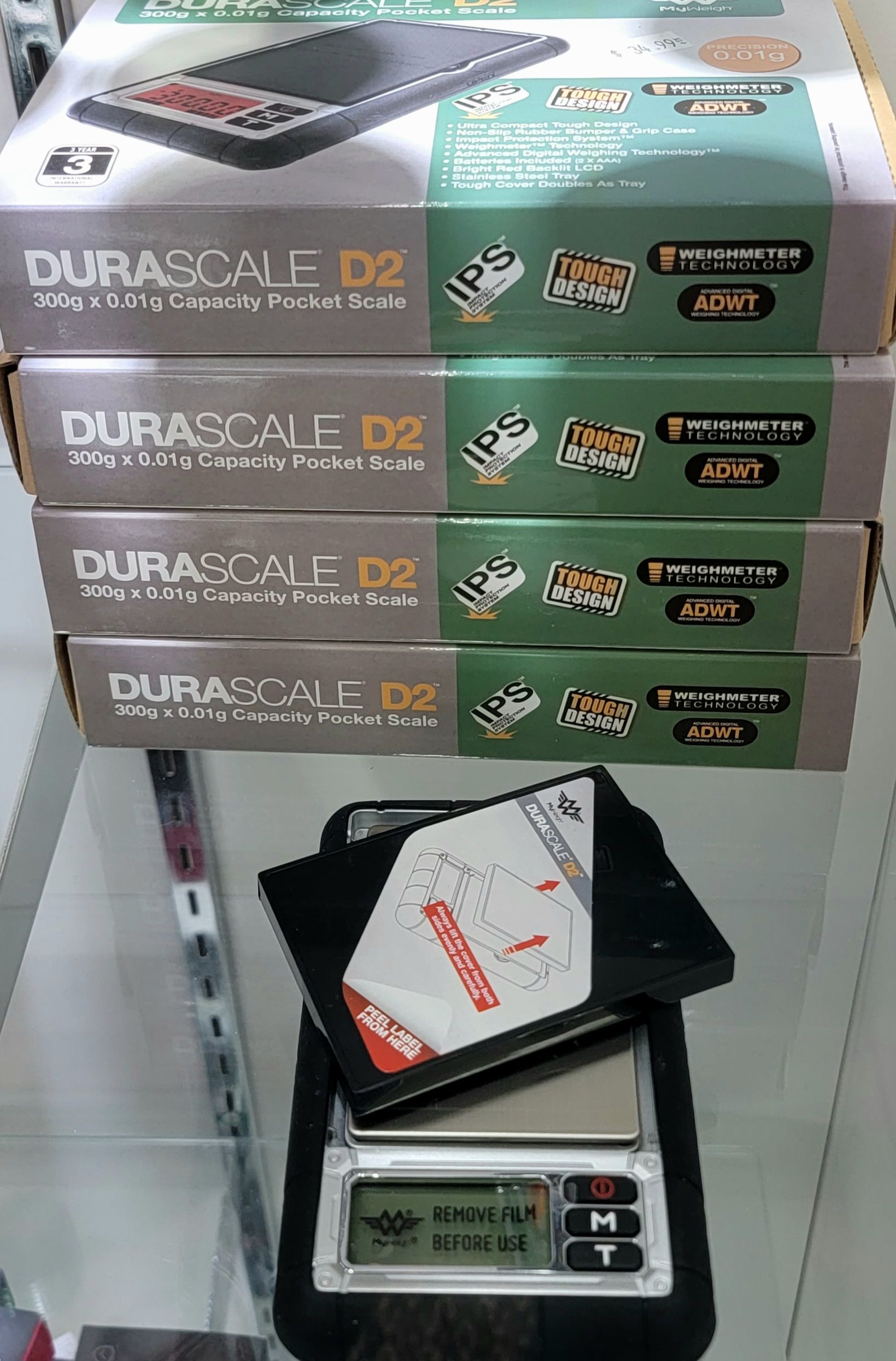 Four boxes of DuraScale D2 pocket scales stacked behind a digital scale, all on a shelf.