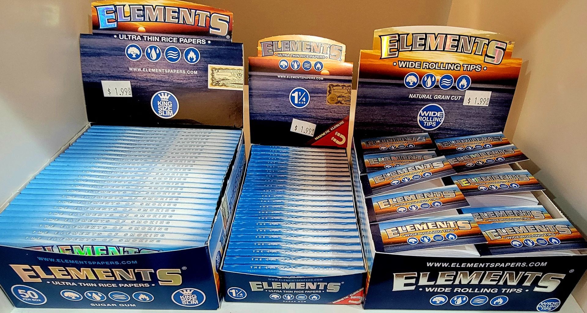 Rolling papers: Elements brand boxes, blue & white, with a sunset background, stacked on a shelf.