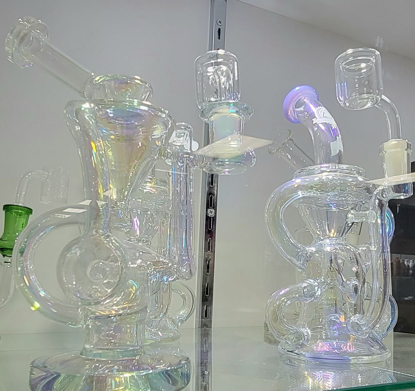 Two clear iridescent glass water pipes on a shelf; one with a green stem, the other with a purple one.