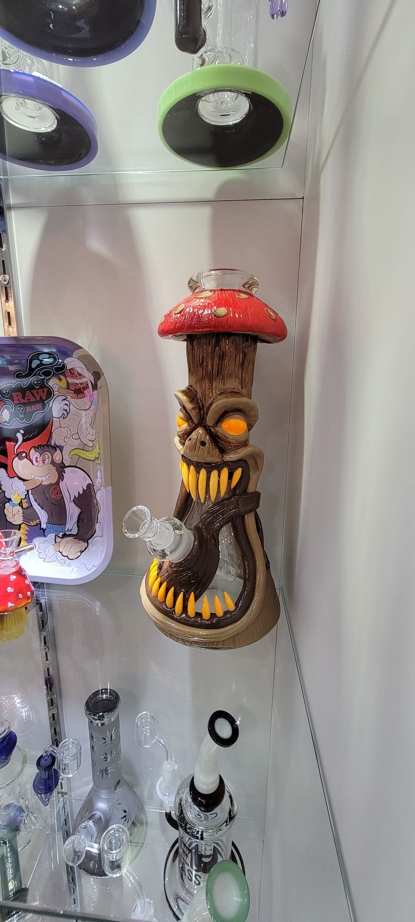 A novelty mushroom-shaped glass bong displayed in a glass cabinet. It has a red cap, brown body, and a toothy grin.