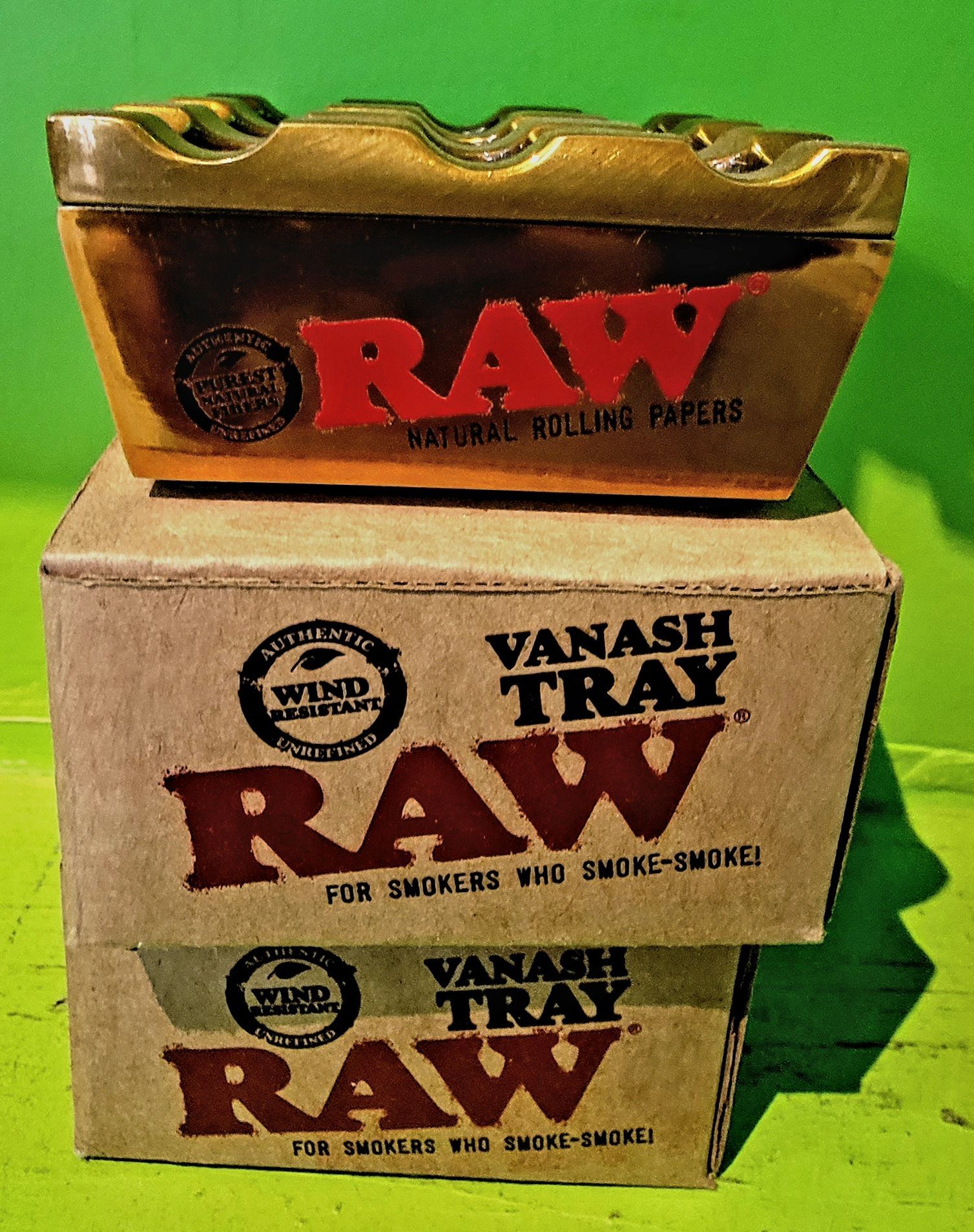 A brown RAW ashtray sits on two brown RAW Vanash Tray boxes. The text is red. Green background.
