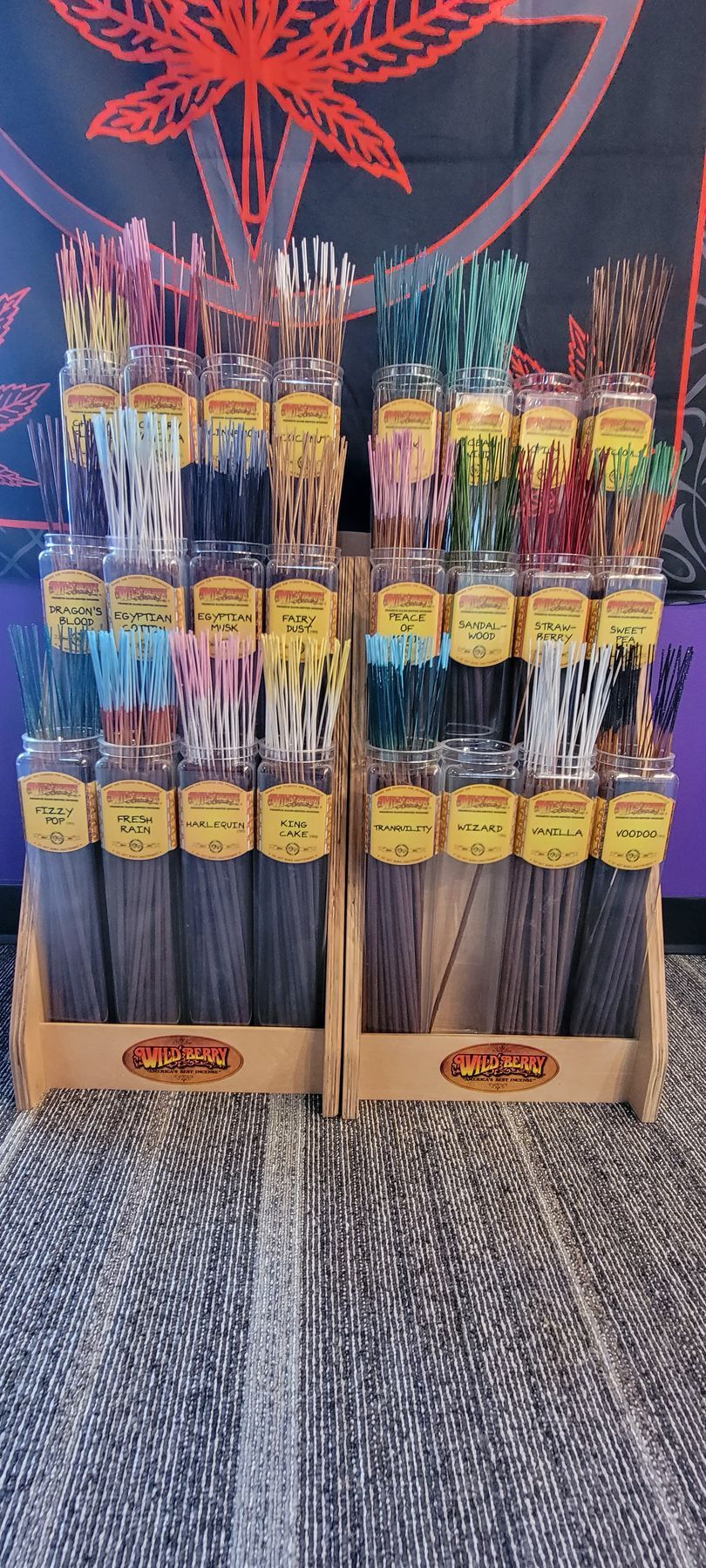 Display of incense sticks in glass jars, arranged in wooden stands. Background has a cannabis leaf design.