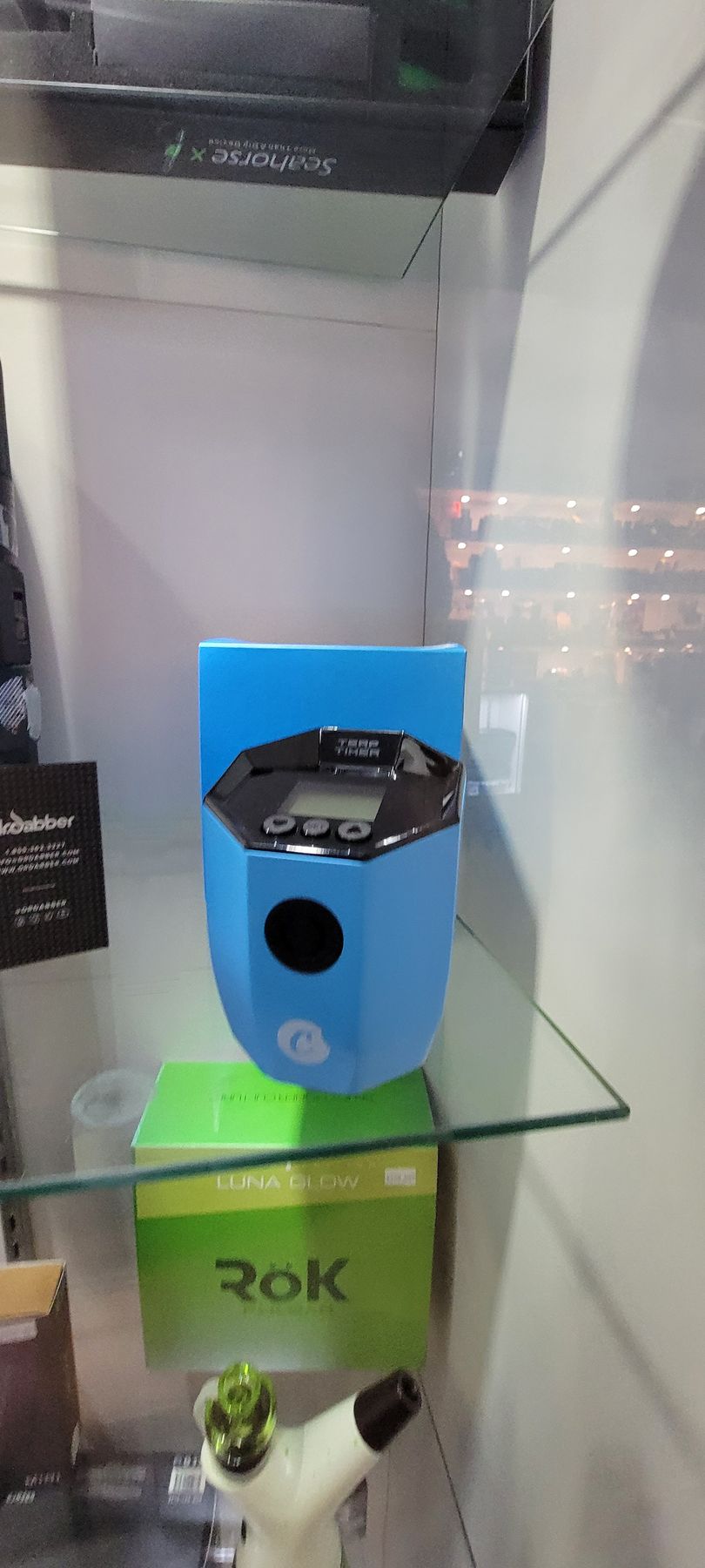 A blue Rok device with a black circle, a green box, and a glass shelf inside a display case.