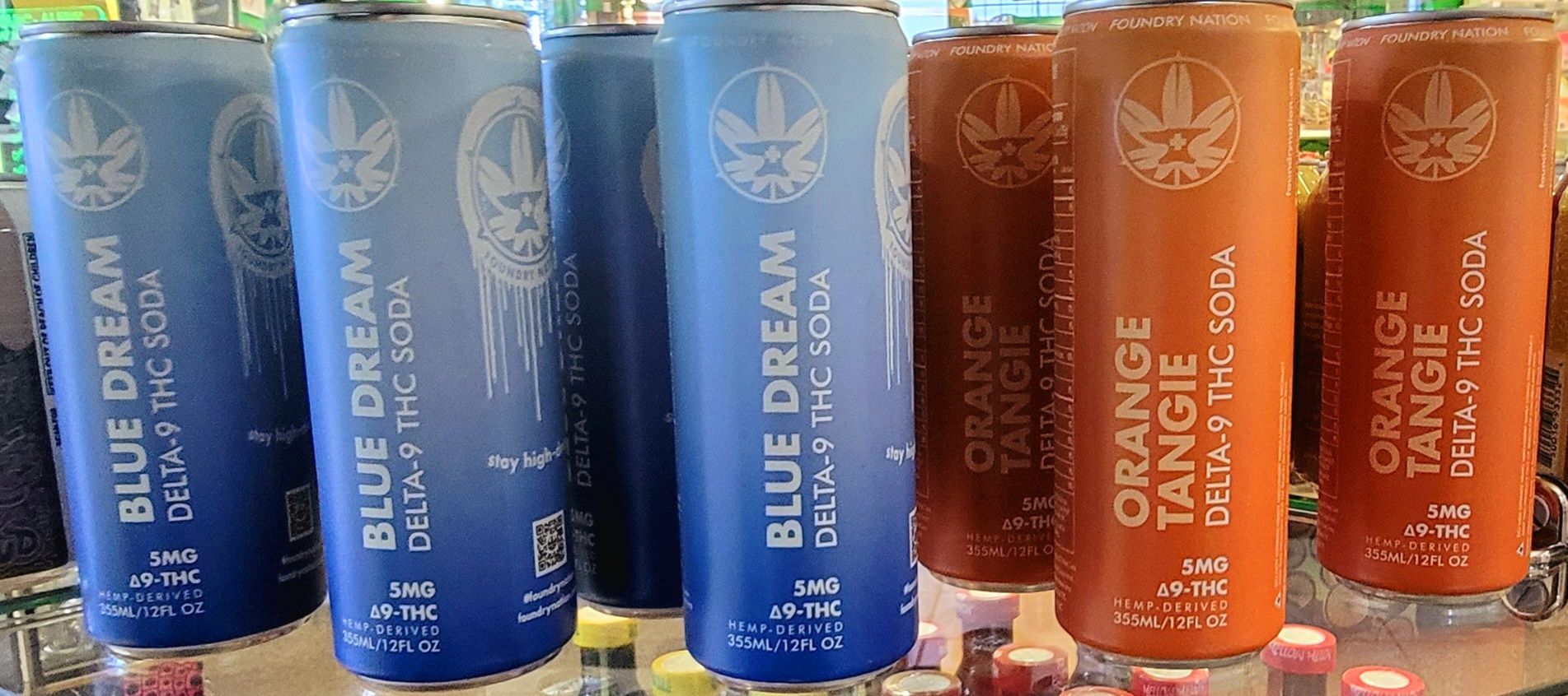 Cans of Blue Dream and Orange Tangie Delta-9 THC soda on a shelf. Blue and orange cans, cannabis leaf logo.