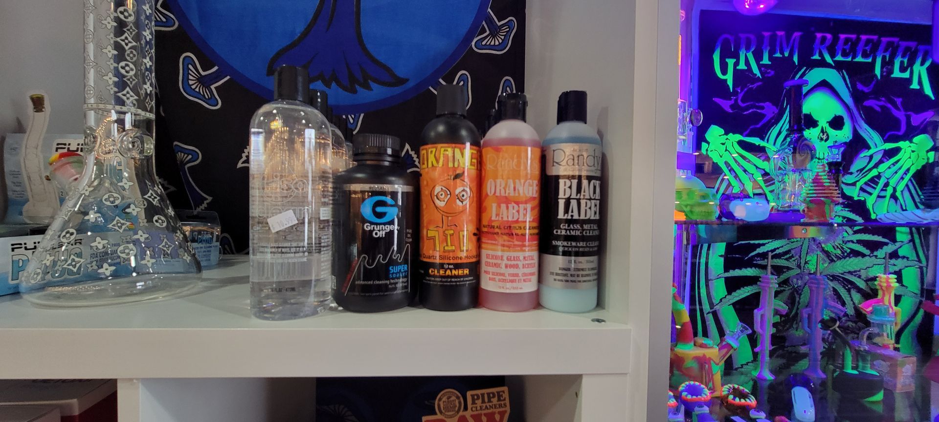 Shelf with various bottles and a water pipe in front of a blue and black poster, and a glow in the dark art piece.