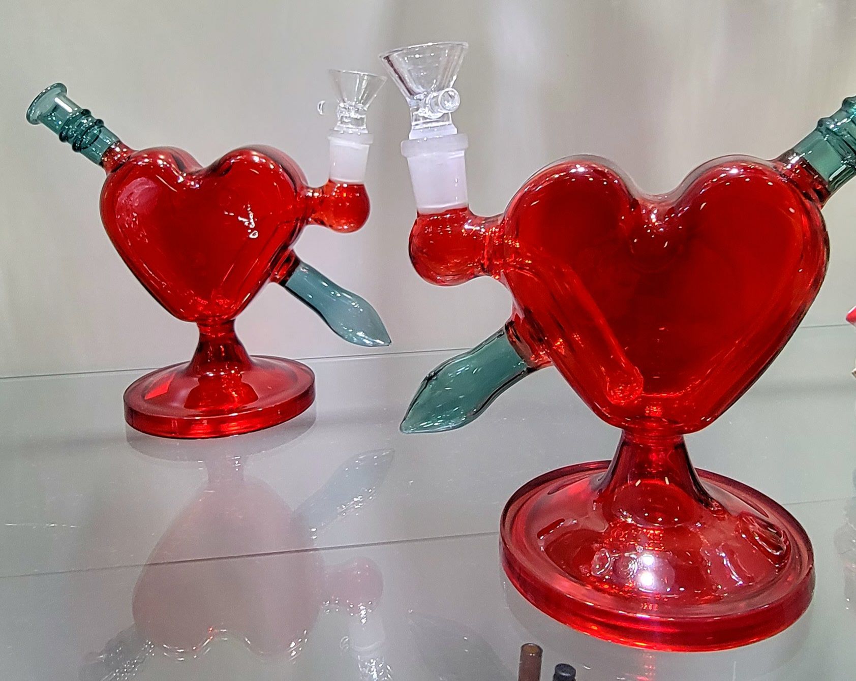 Two red heart-shaped glass bongs with a green arrow through them, resting on a reflective surface.