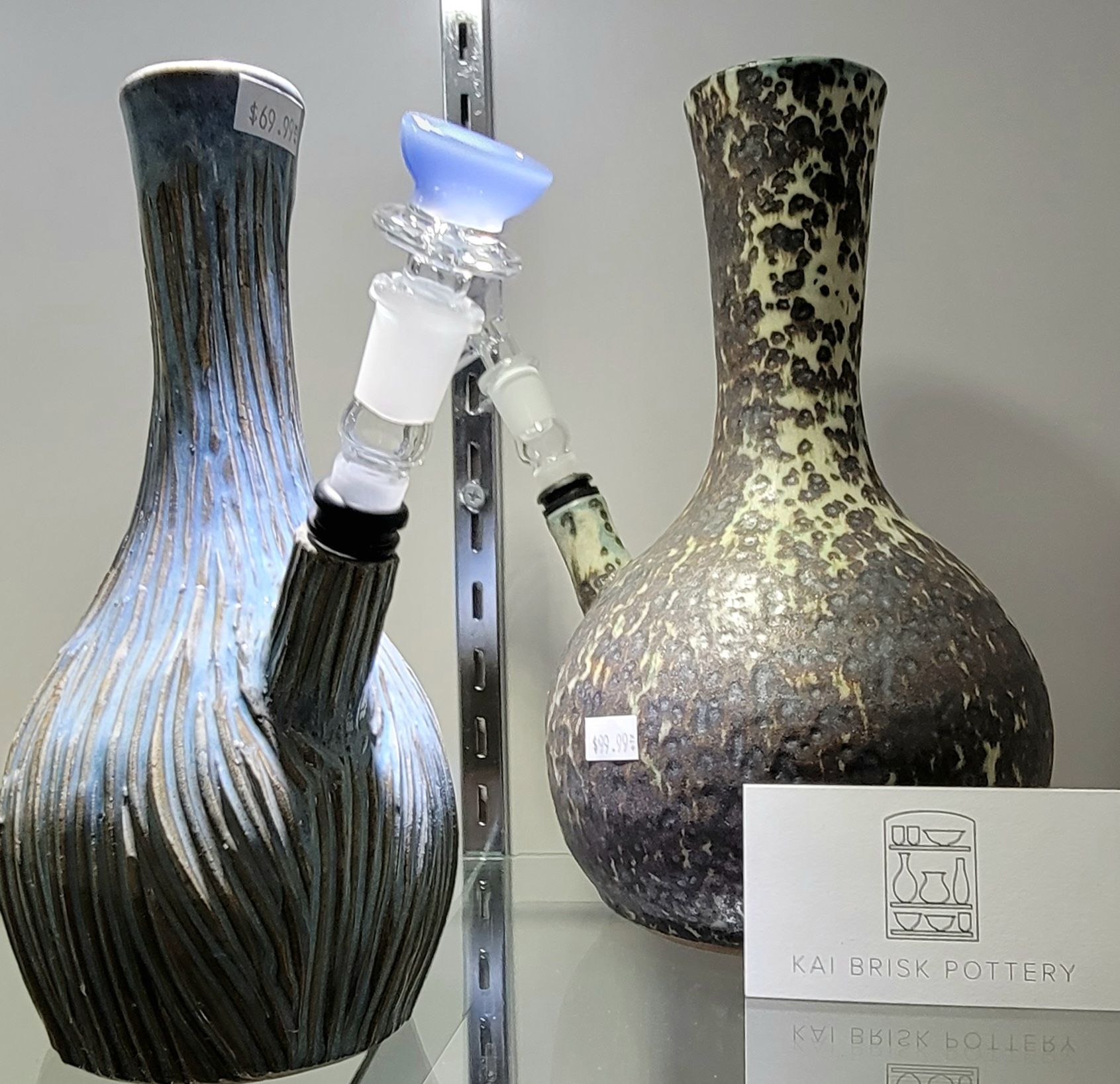 Two ceramic bongs with glass attachments, on a shelf. One is blue, the other brown/yellow. A business card sits in front.