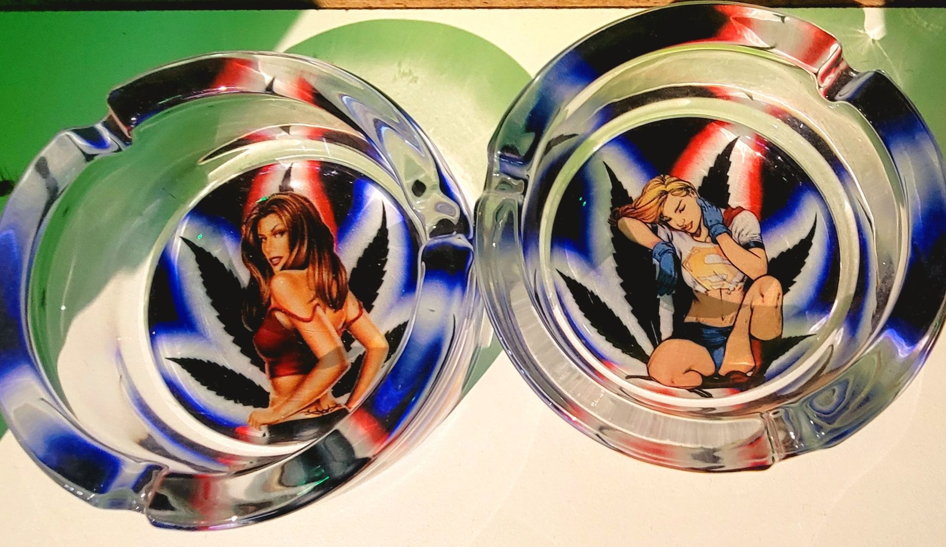 Two clear glass ashtrays with women posing, set against a cannabis leaf design with red, white, and blue.