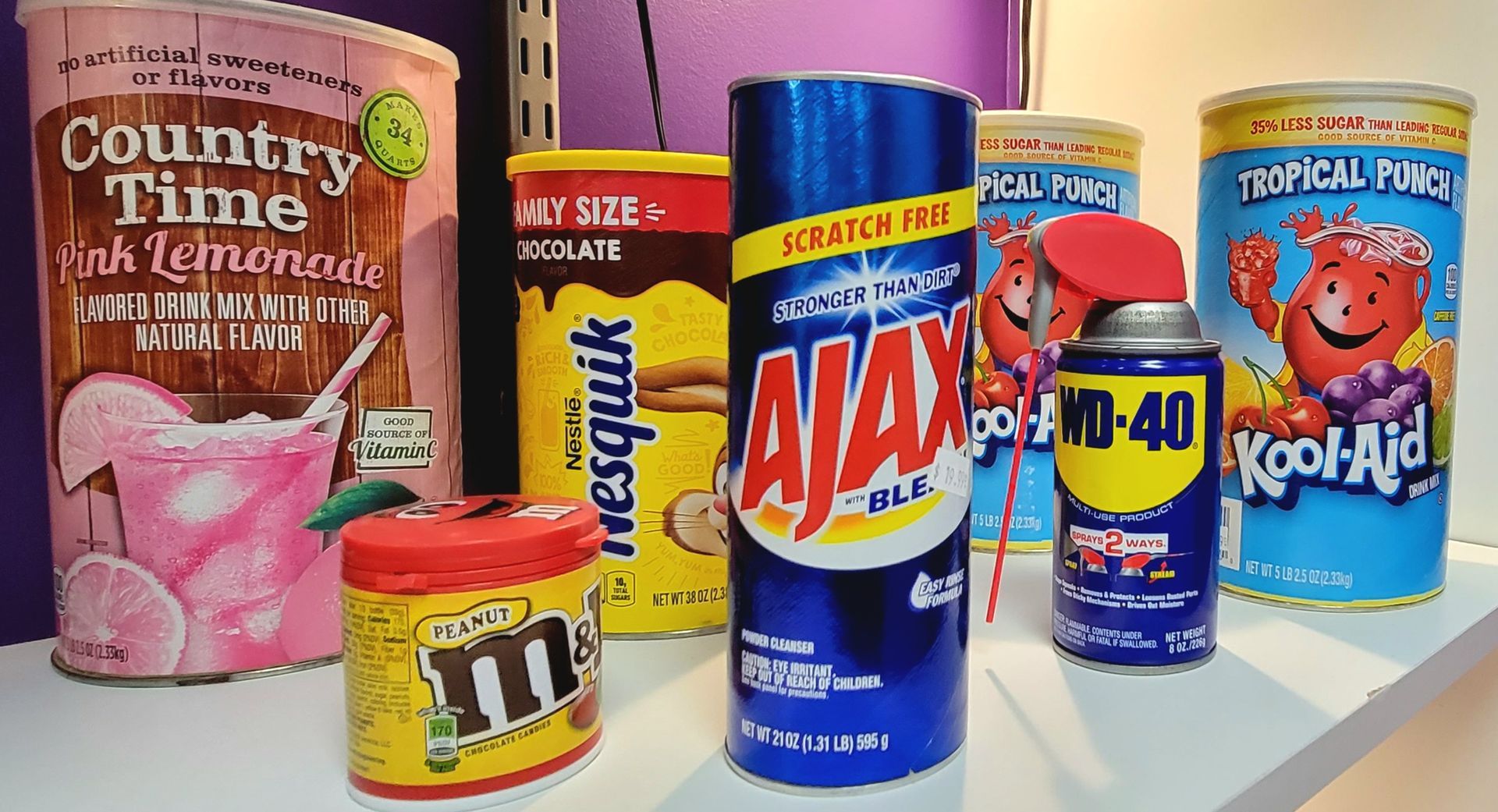 Assortment of product containers like lemonade, cocoa, Ajax, WD-40, M&Ms, and Kool-Aid displayed on a shelf.