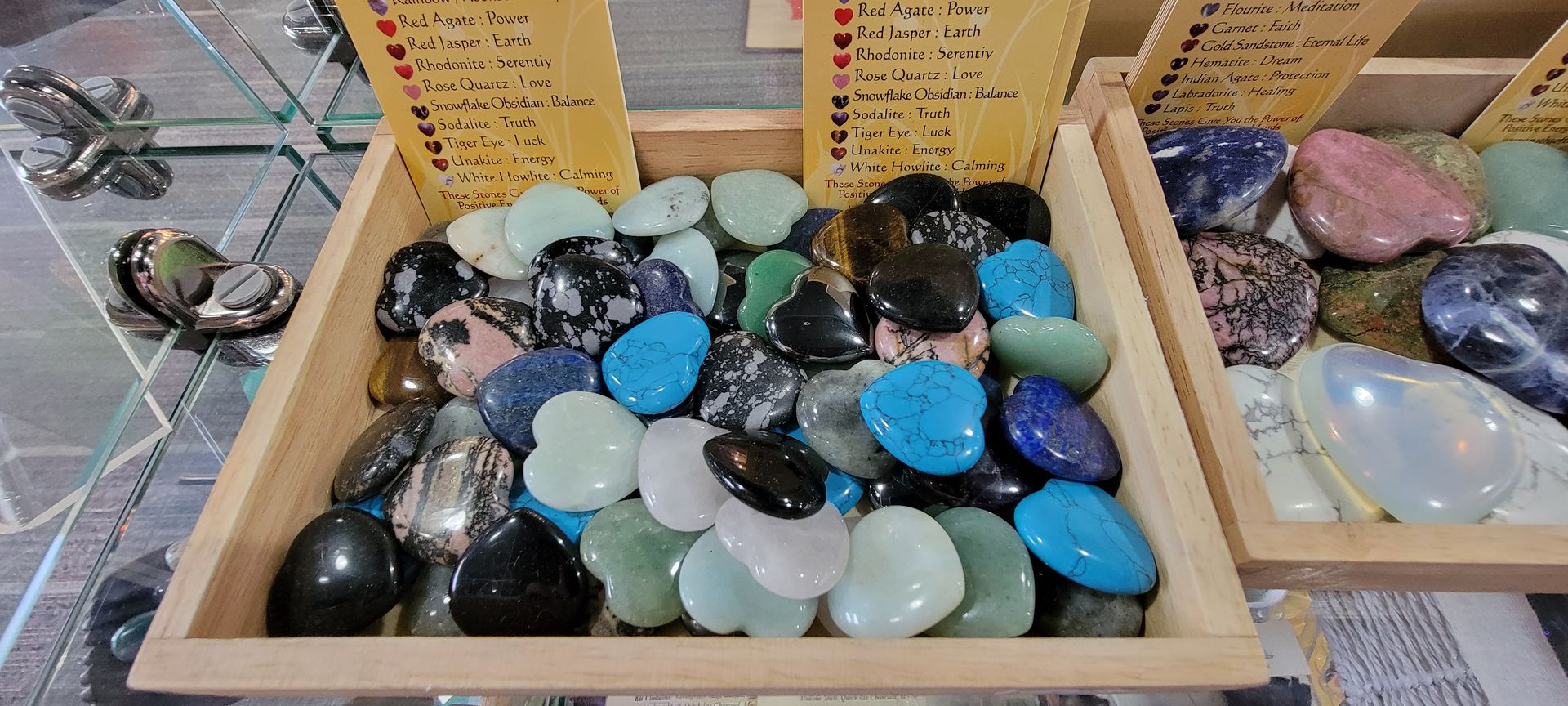 Wooden trays filled with colorful heart-shaped polished gemstones, on a reflective surface. Signs list the stones.